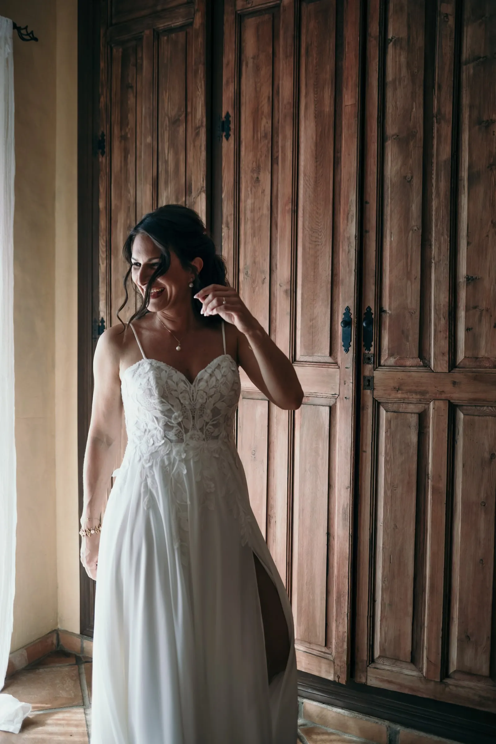 La novia, con vestido de encaje, sonríe junto a una gran puerta de madera mientras se prepara, Hacienda San José, Mijas