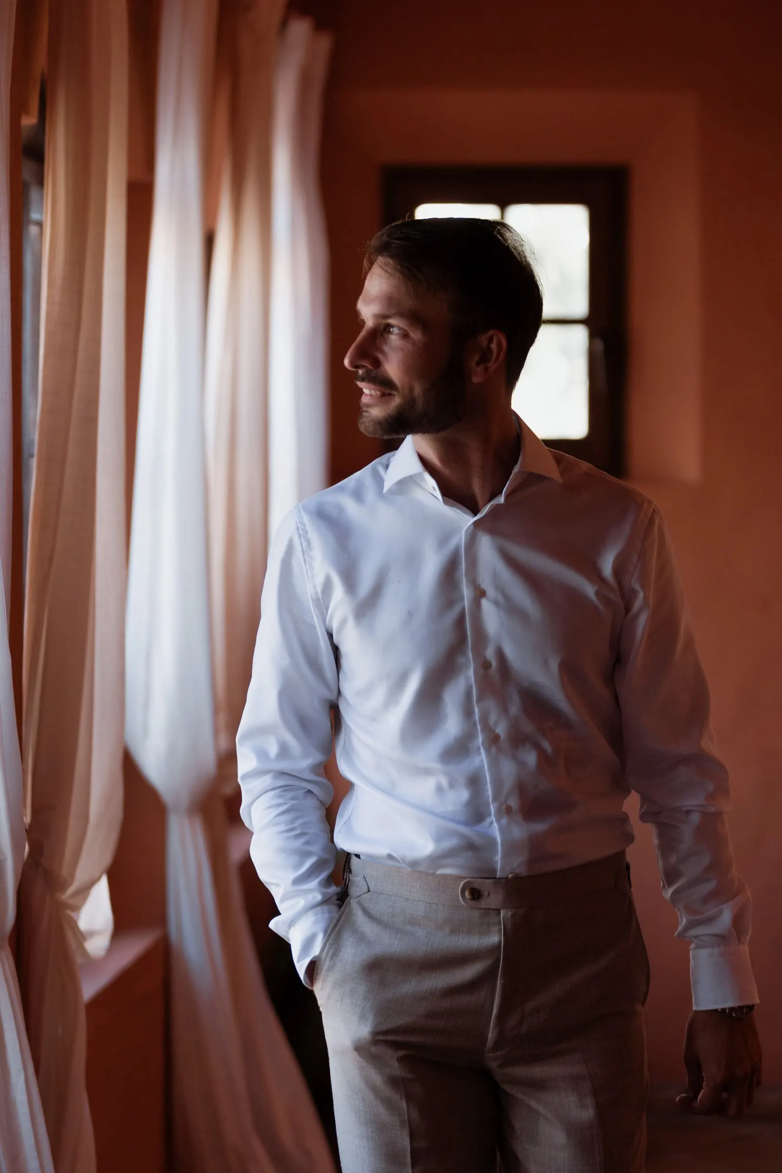 El novio, con camisa blanca, sonríe junto a una ventana con cortinas en una sala color teja, Hacienda San José, Mijas