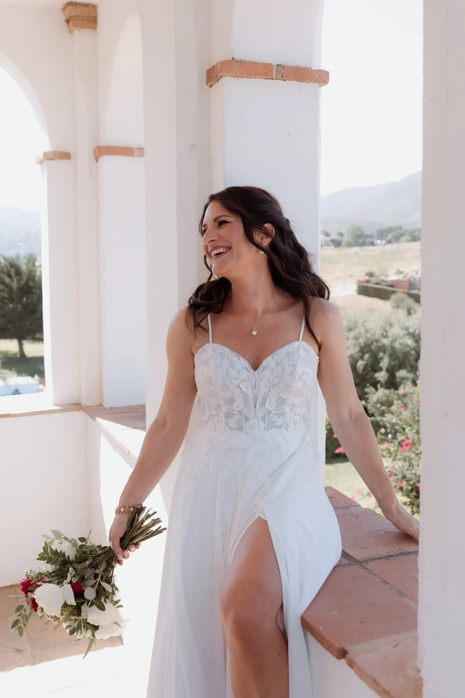 La novia se ríe en una terraza encalada con su ramo de rosas rojas y blancas, Hacienda San José, Mijas
