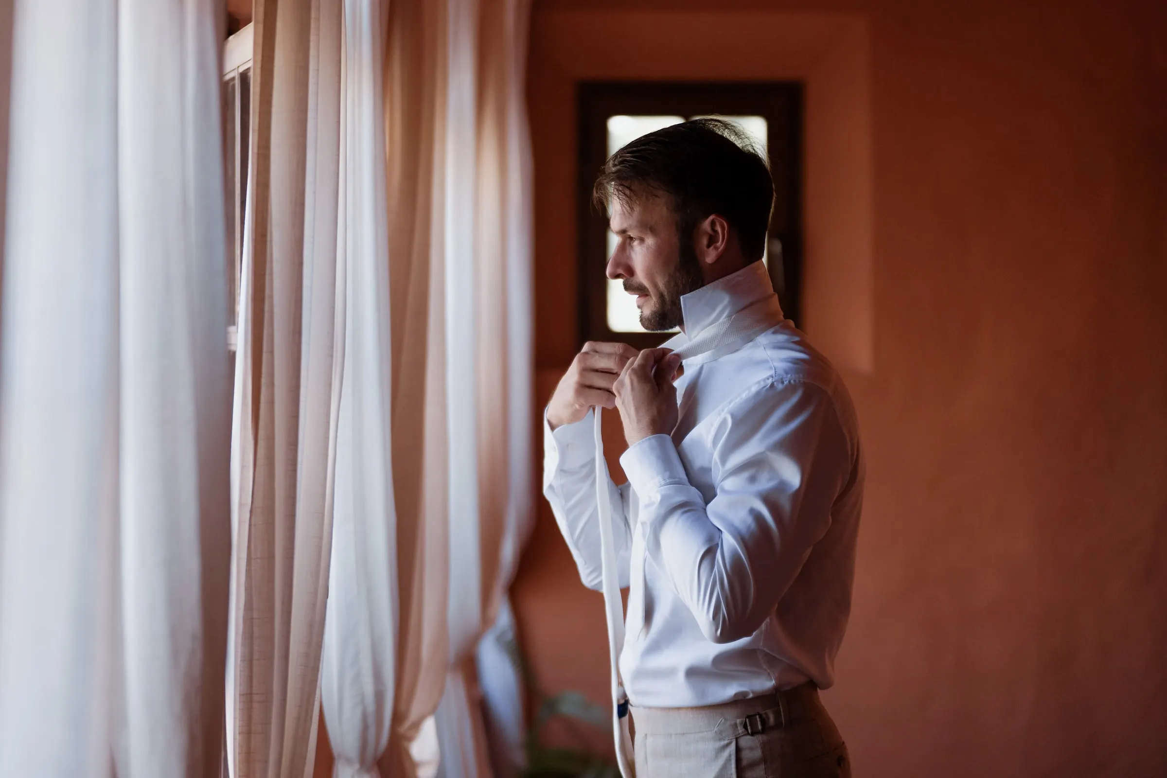 El novio se abrocha el cuello de la camisa junto a una ventana con cortinas, de perfil, Hacienda San José, Mijas