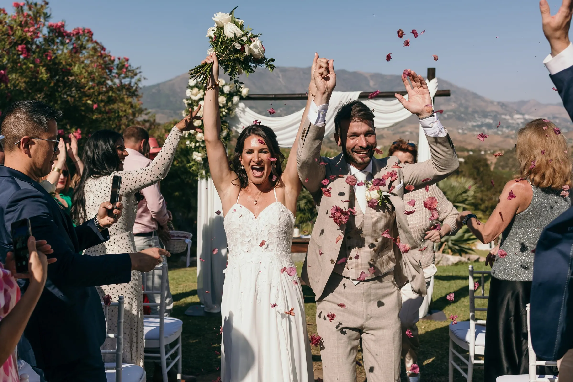 Los novios celebran con los brazos en alto mientras los invitados lanzan pétalos rojos, Hacienda San José, Mijas