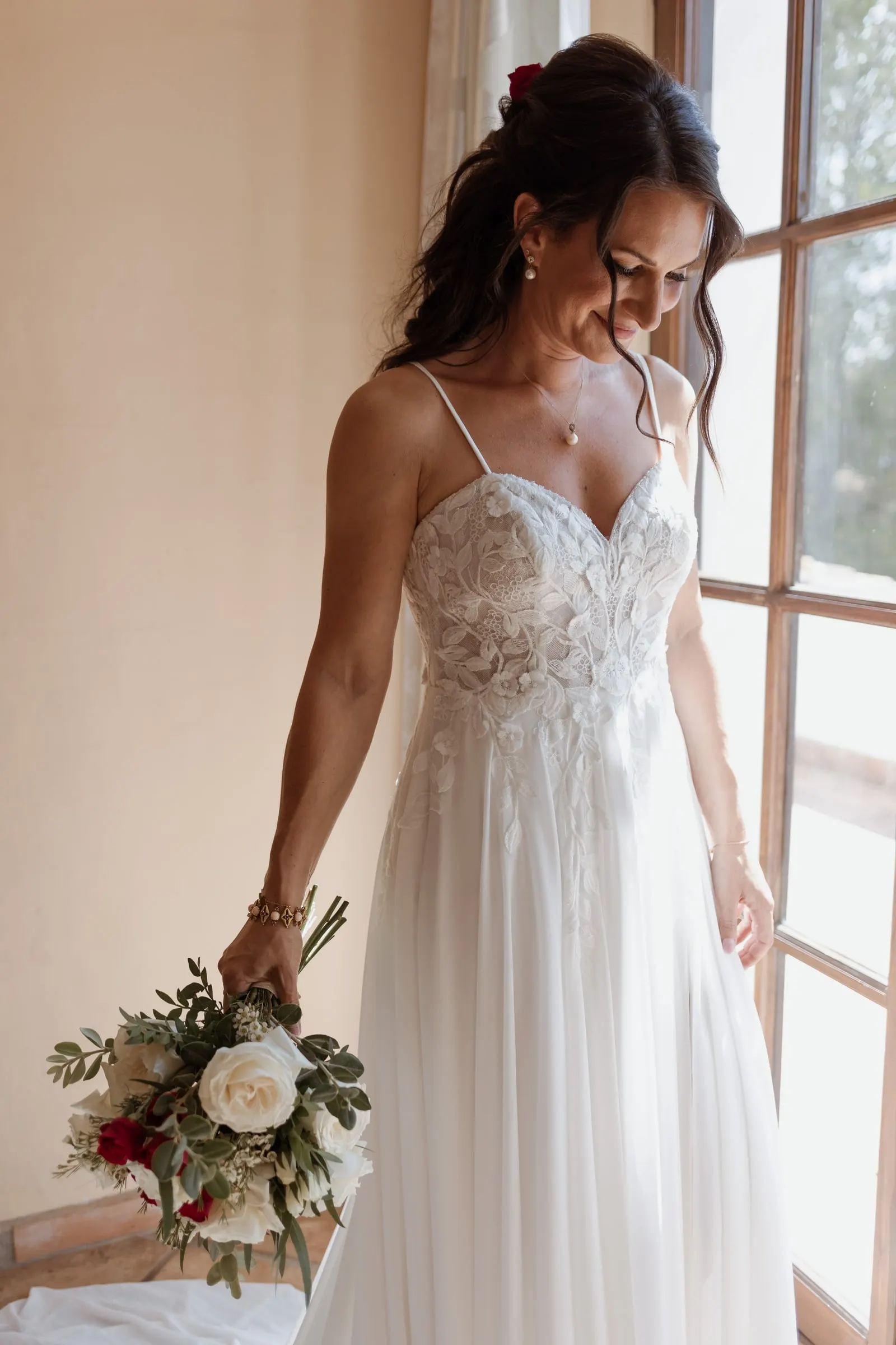 La novia mira su vestido junto a un gran ventanal, con el ramo en la mano, Hacienda San José, Mijas