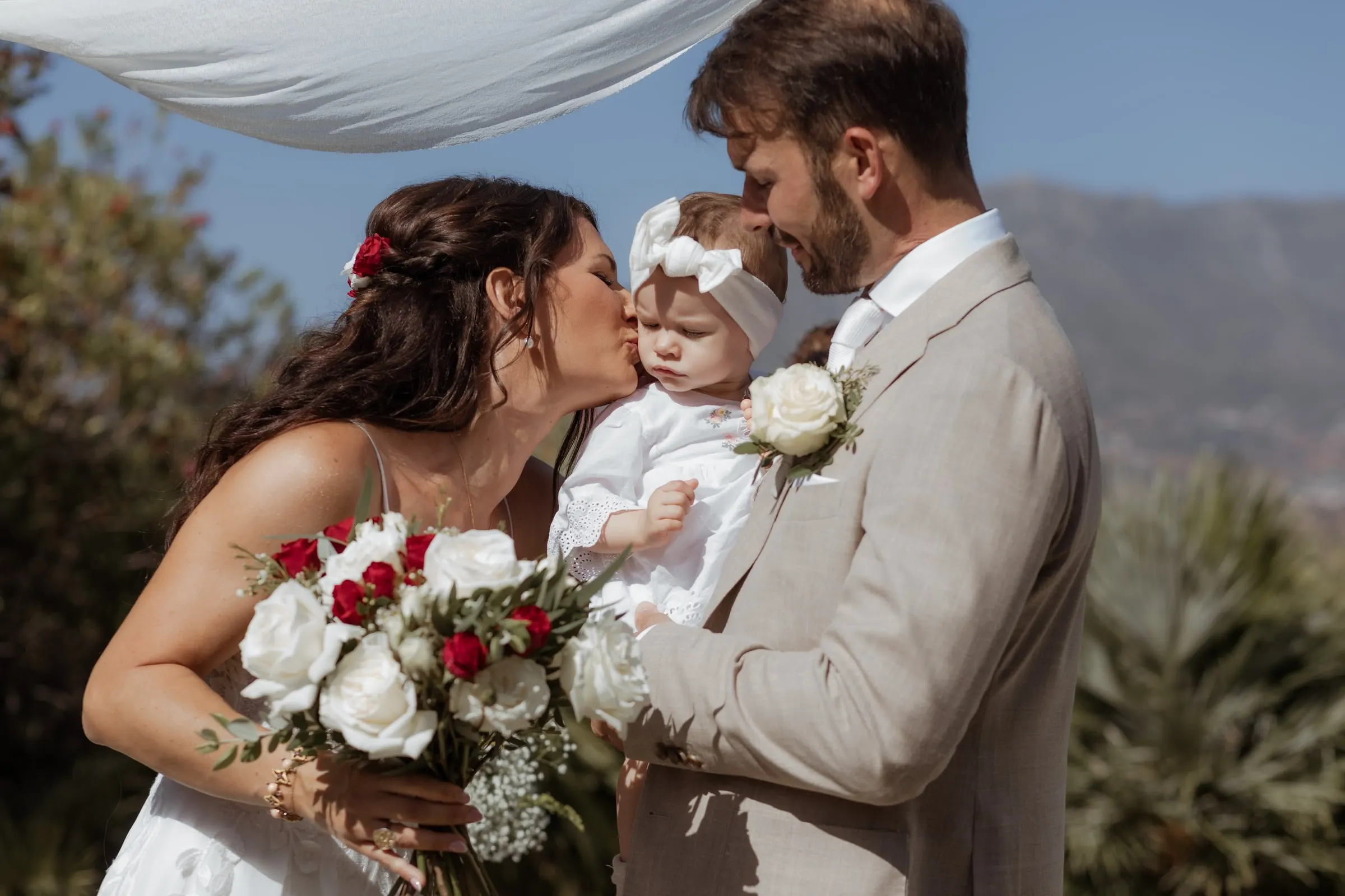 Los recién casados besan a su bebé, con lazo blanco, bajo el dosel de la ceremonia, Hacienda San José, Mijas