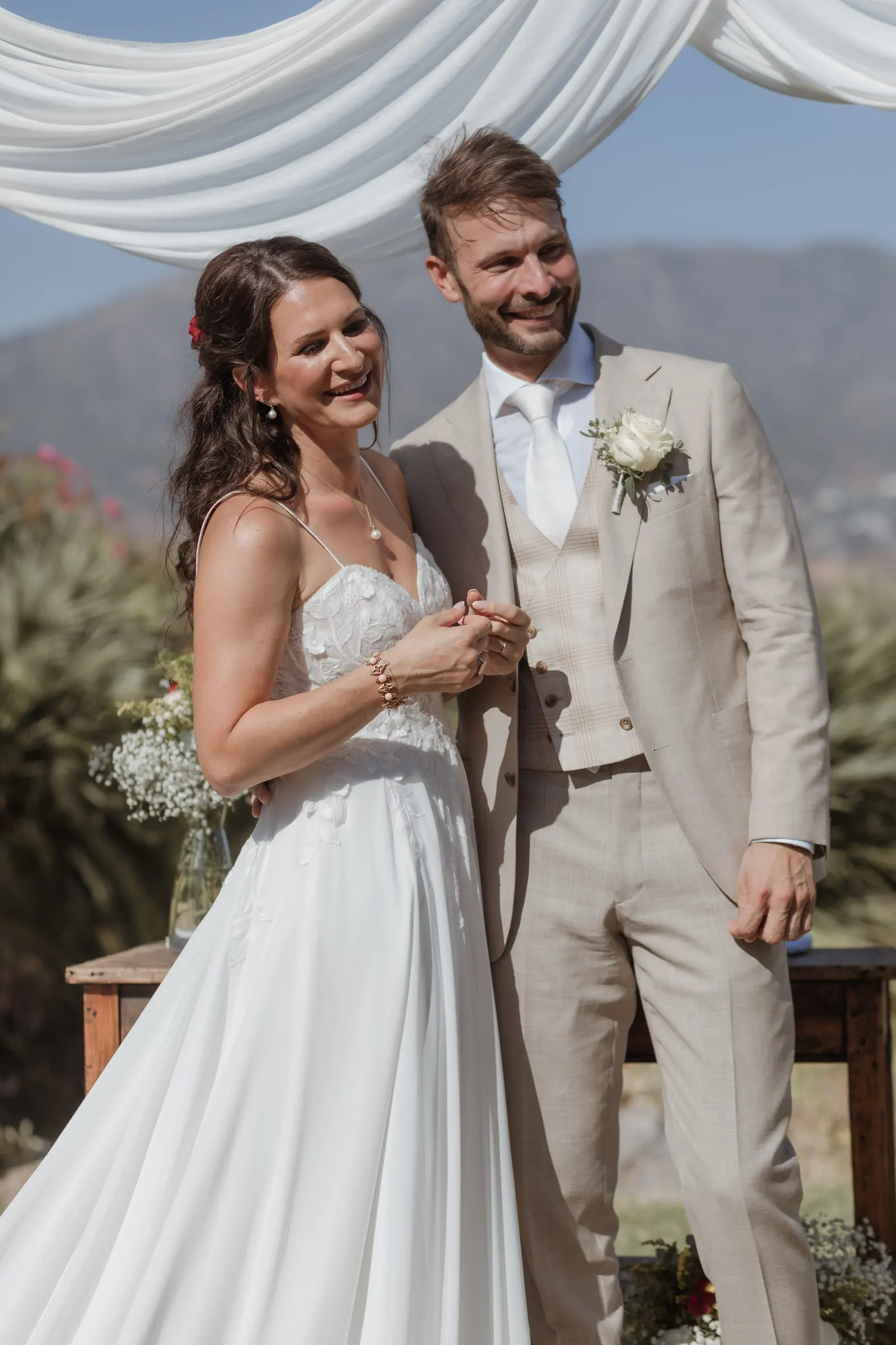 Los novios se ríen uno junto al otro en la mesa de ceremonia, con la sierra detrás, Hacienda San José, Mijas