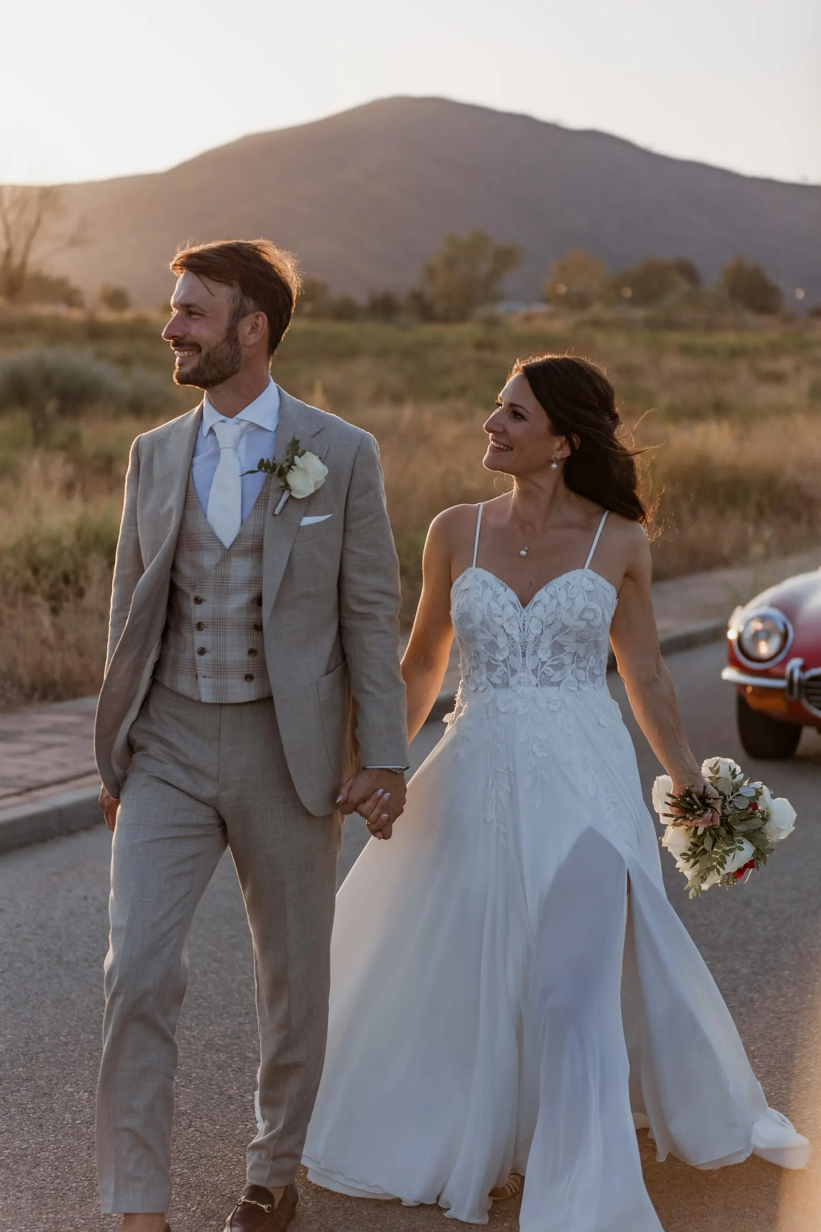 Los novios caminan de la mano por un camino al atardecer con un clásico rojo detrás, Hacienda San José, Mijas