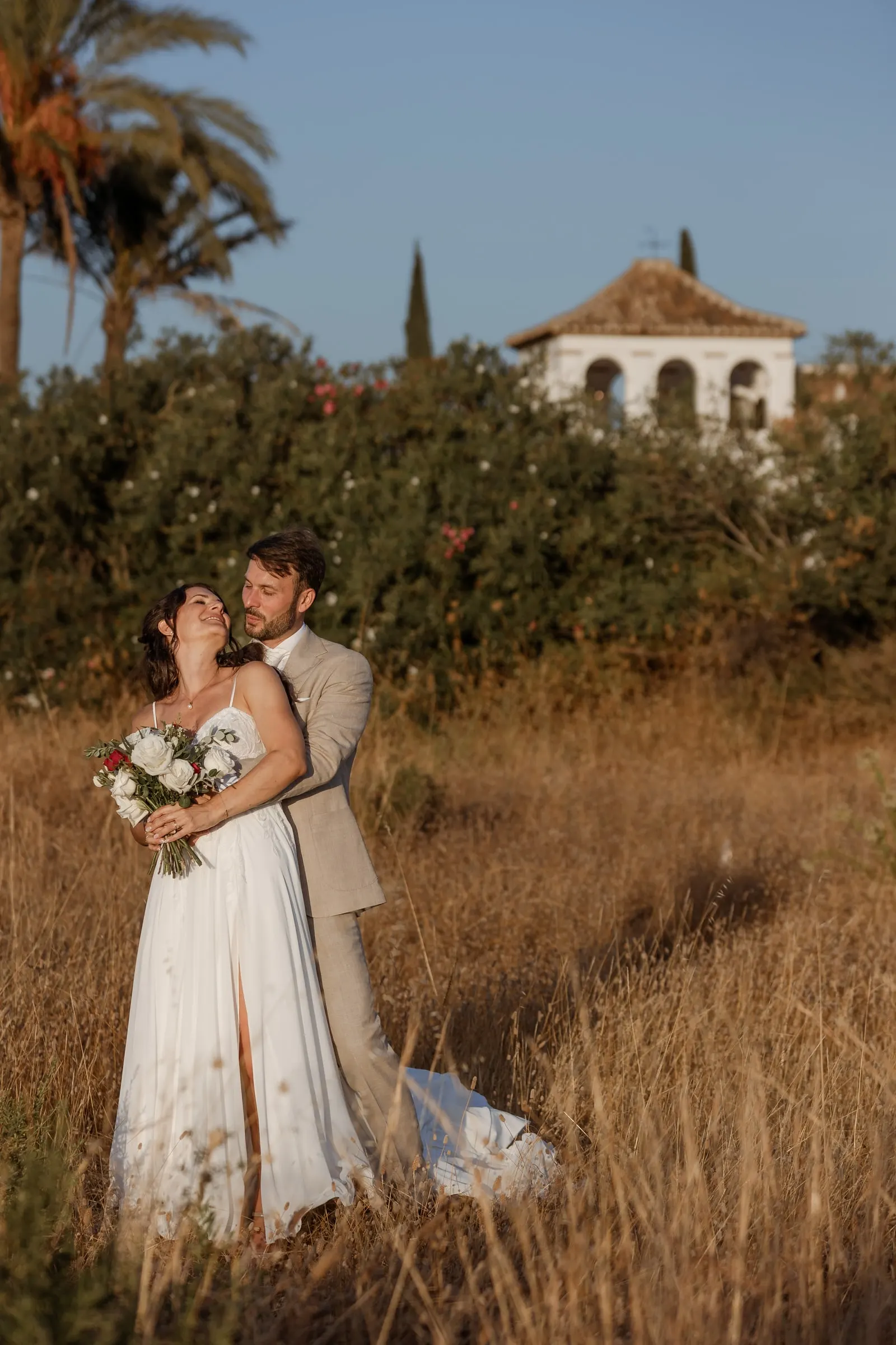 Los novios se abrazan entre la hierba alta y dorada con el campanario detrás, Hacienda San José, Mijas