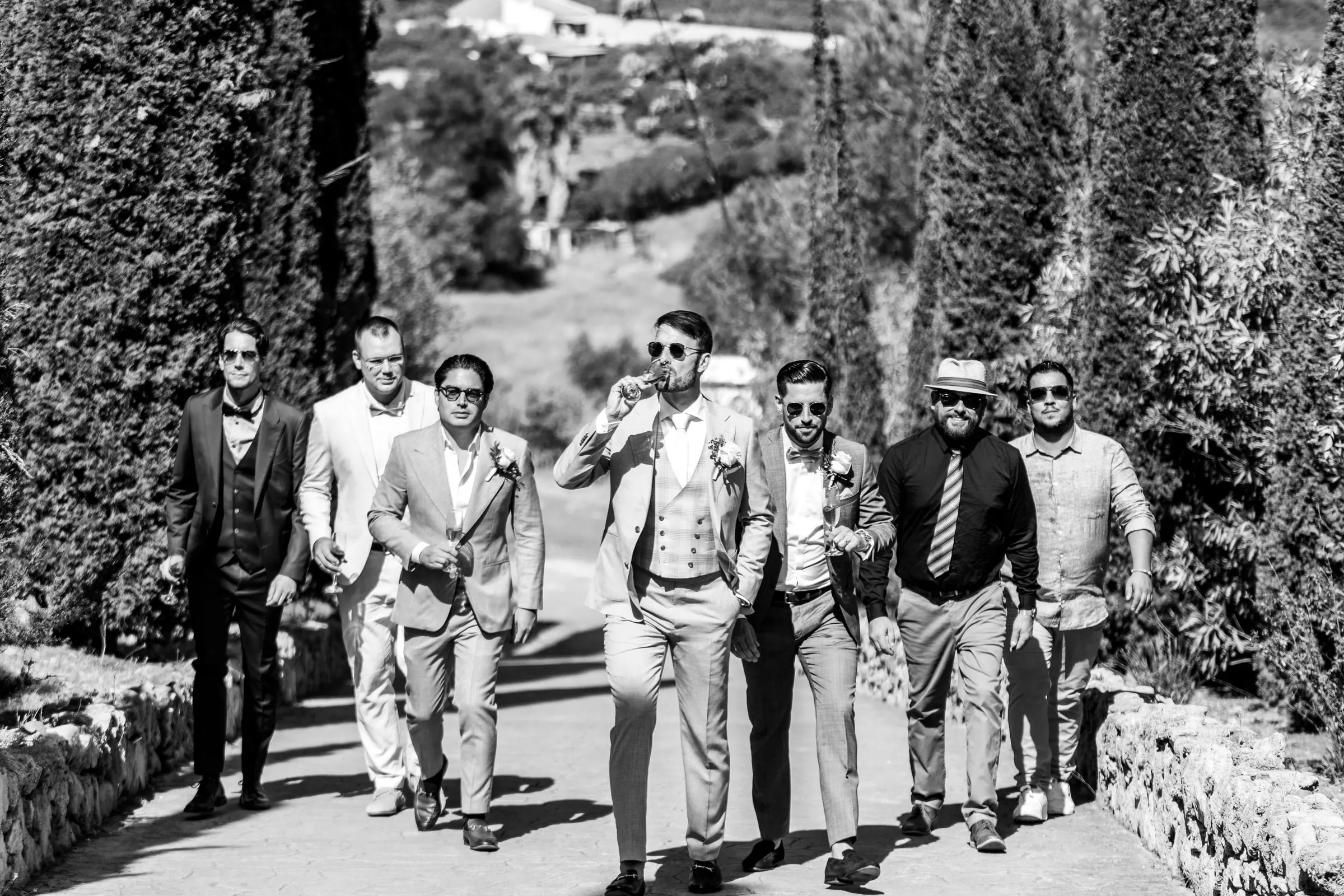 El novio y sus amigos suben con gafas de sol por un camino arbolado, en blanco y negro, Hacienda San José, Mijas