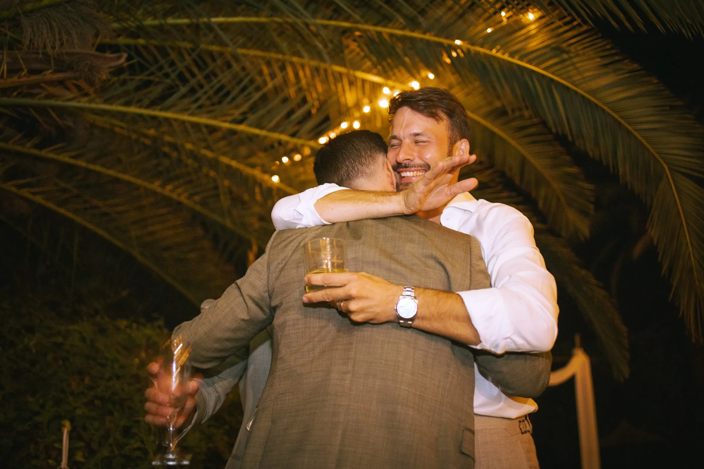 Un amigo abraza al novio bajo las hojas de palmera y las guirnaldas de luces, Hacienda San José, Mijas