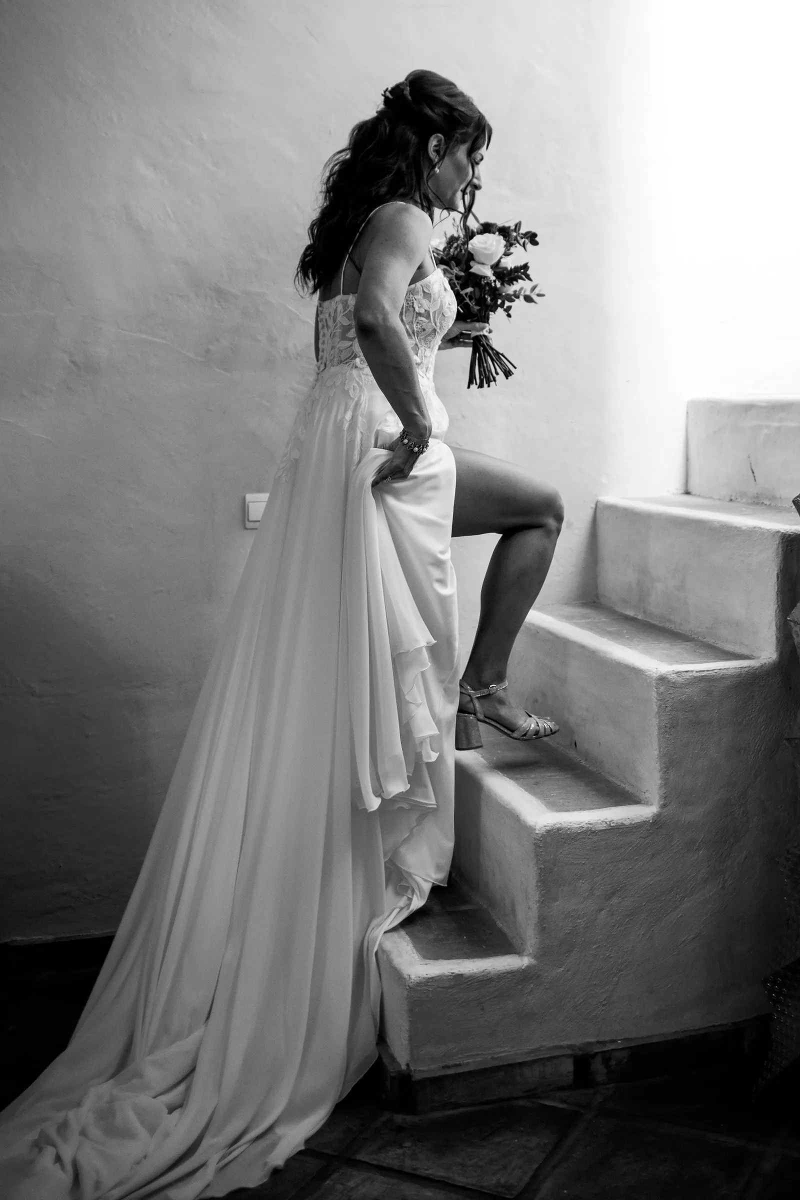 La novia sube unos escalones blancos oliendo su ramo, en blanco y negro, Hacienda San José, Mijas