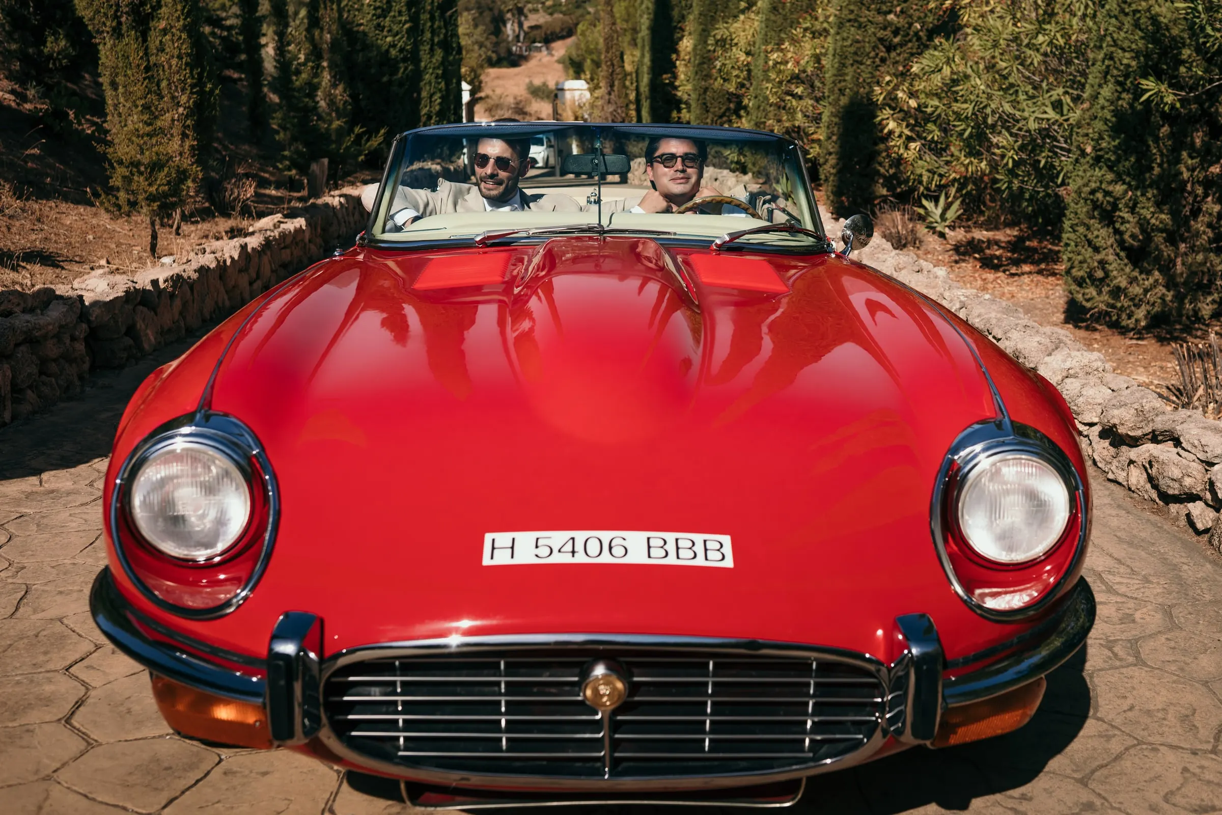 Dos hombres al volante de un descapotable rojo antiguo en un camino empedrado, de frente, Hacienda San José, Mijas