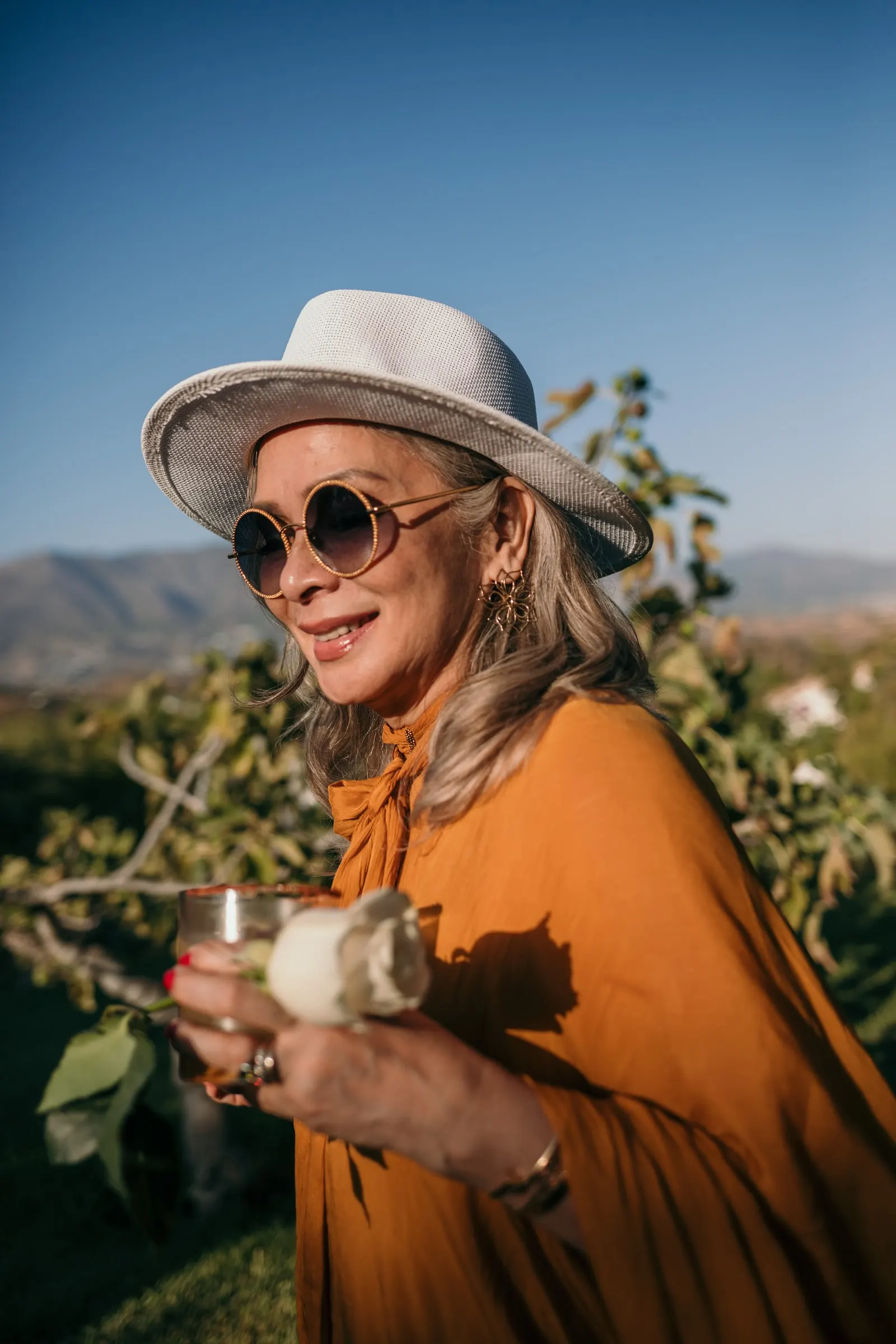 Una invitada con sombrero y gafas redondas sostiene una copa al sol, Hacienda San José, Mijas
