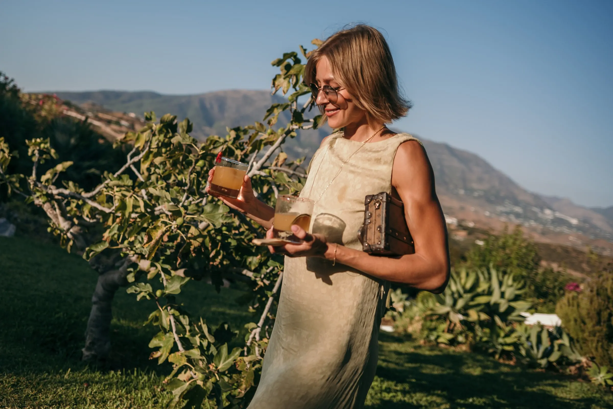 Una invitada con vestido de lino sostiene unas copas junto a una higuera, con la sierra detrás, Hacienda San José, Mijas