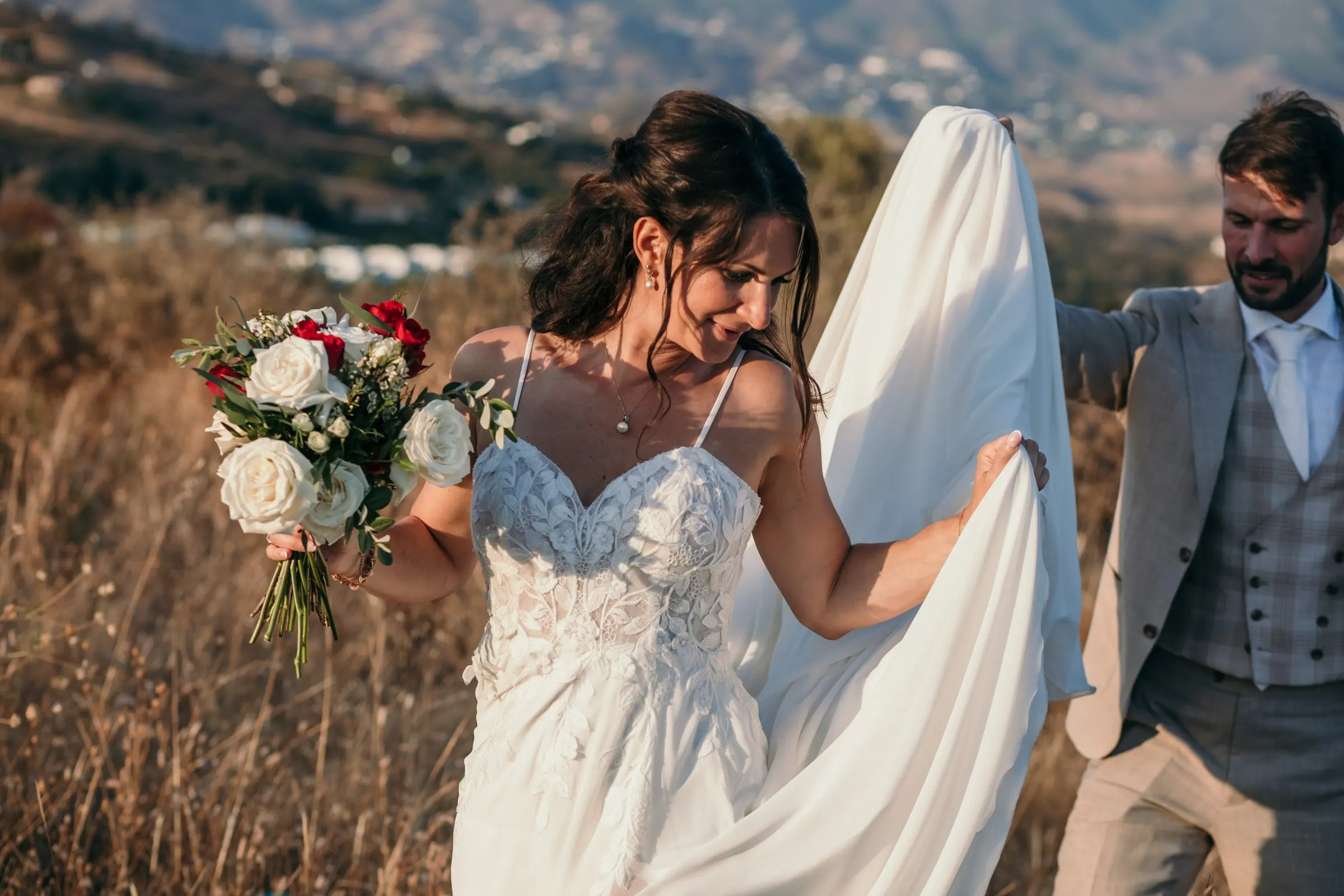 La novia se ríe mientras el novio le levanta la cola del vestido en un campo seco, Hacienda San José, Mijas