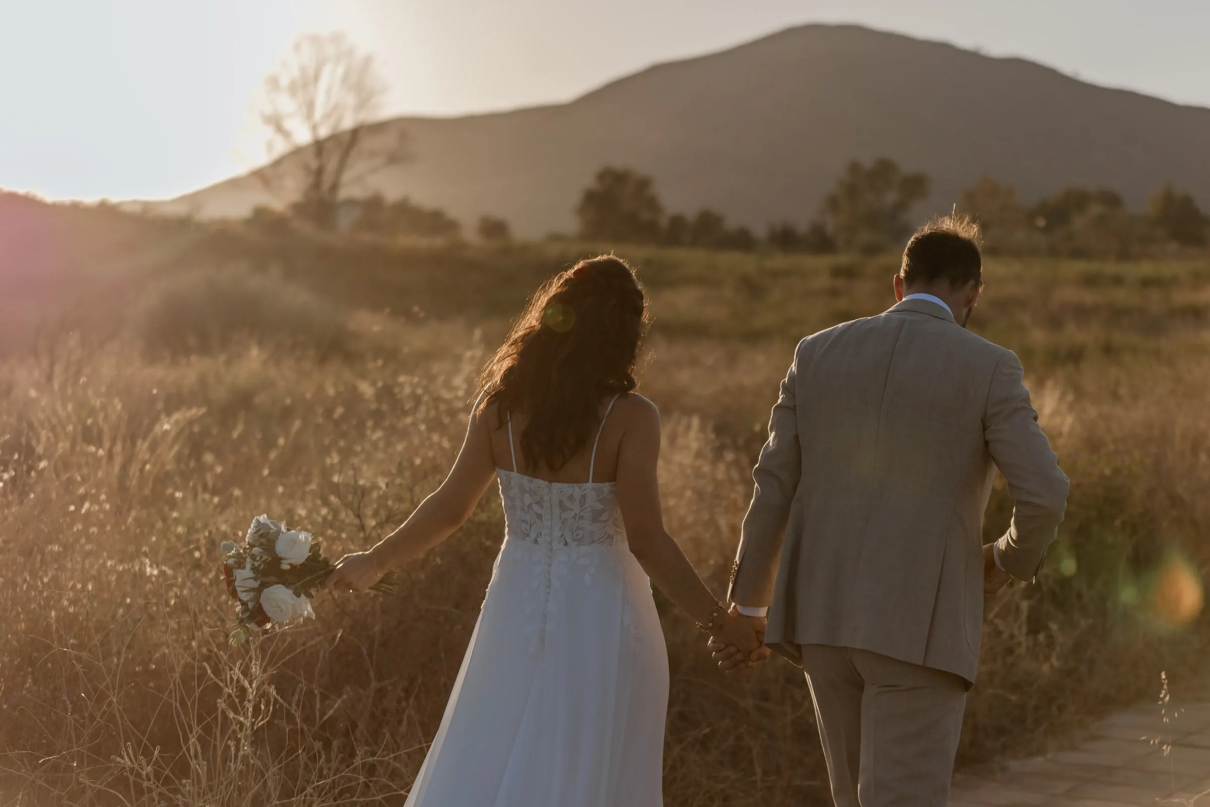 Los novios, de espaldas, caminan de la mano entre la hierba dorada al atardecer, Hacienda San José, Mijas