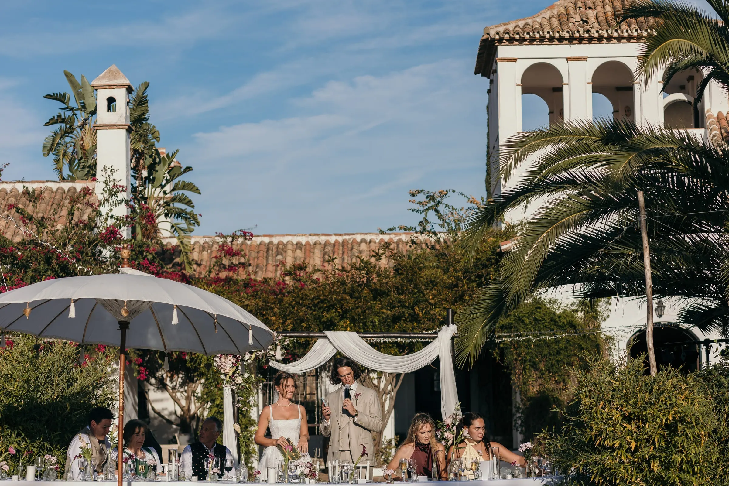 The newlyweds standing for speeches at the head table under white parasols, Hacienda San José, Mijas