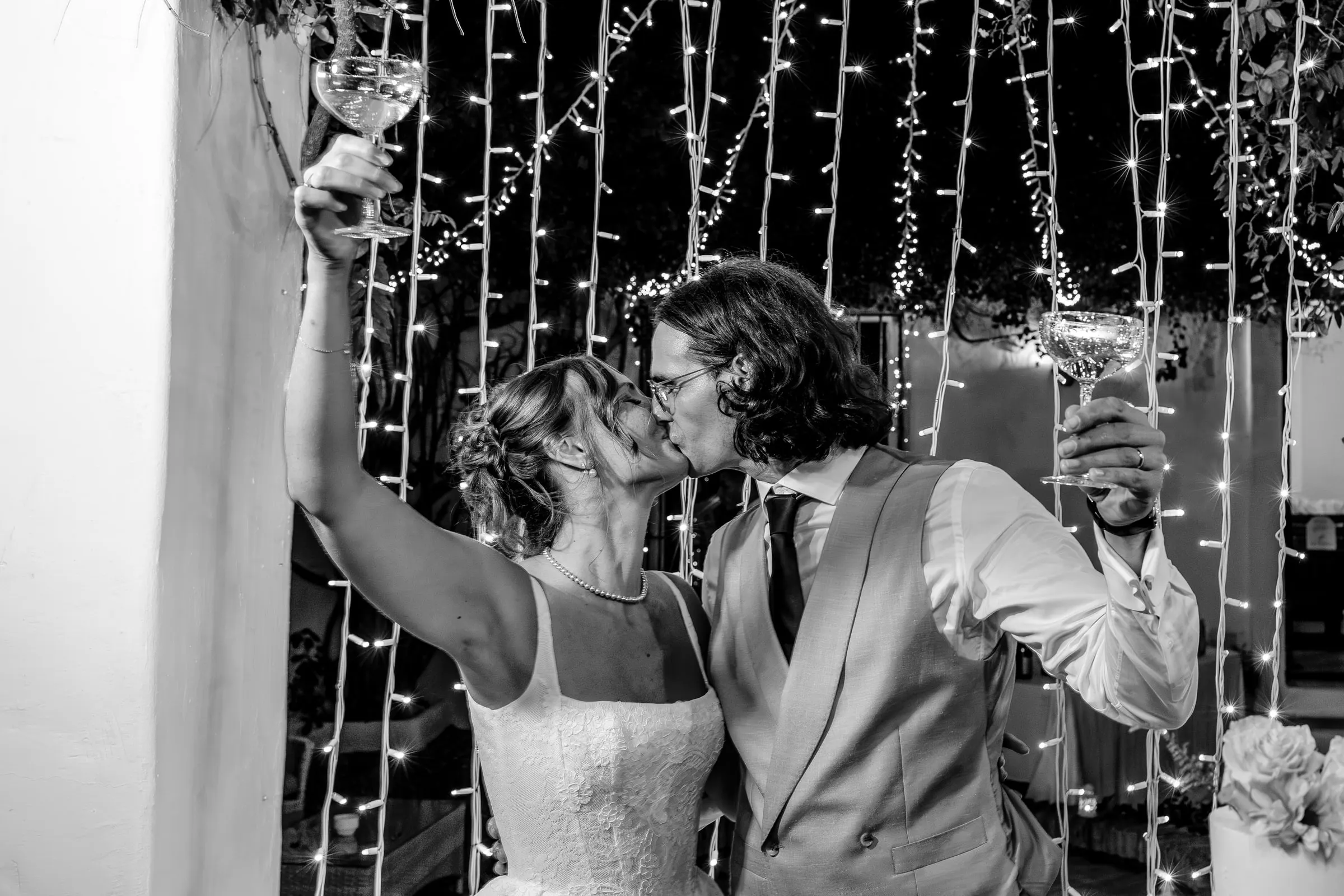 Black and white of the couple kissing with raised glasses before a curtain of lights, Hacienda San José, Mijas