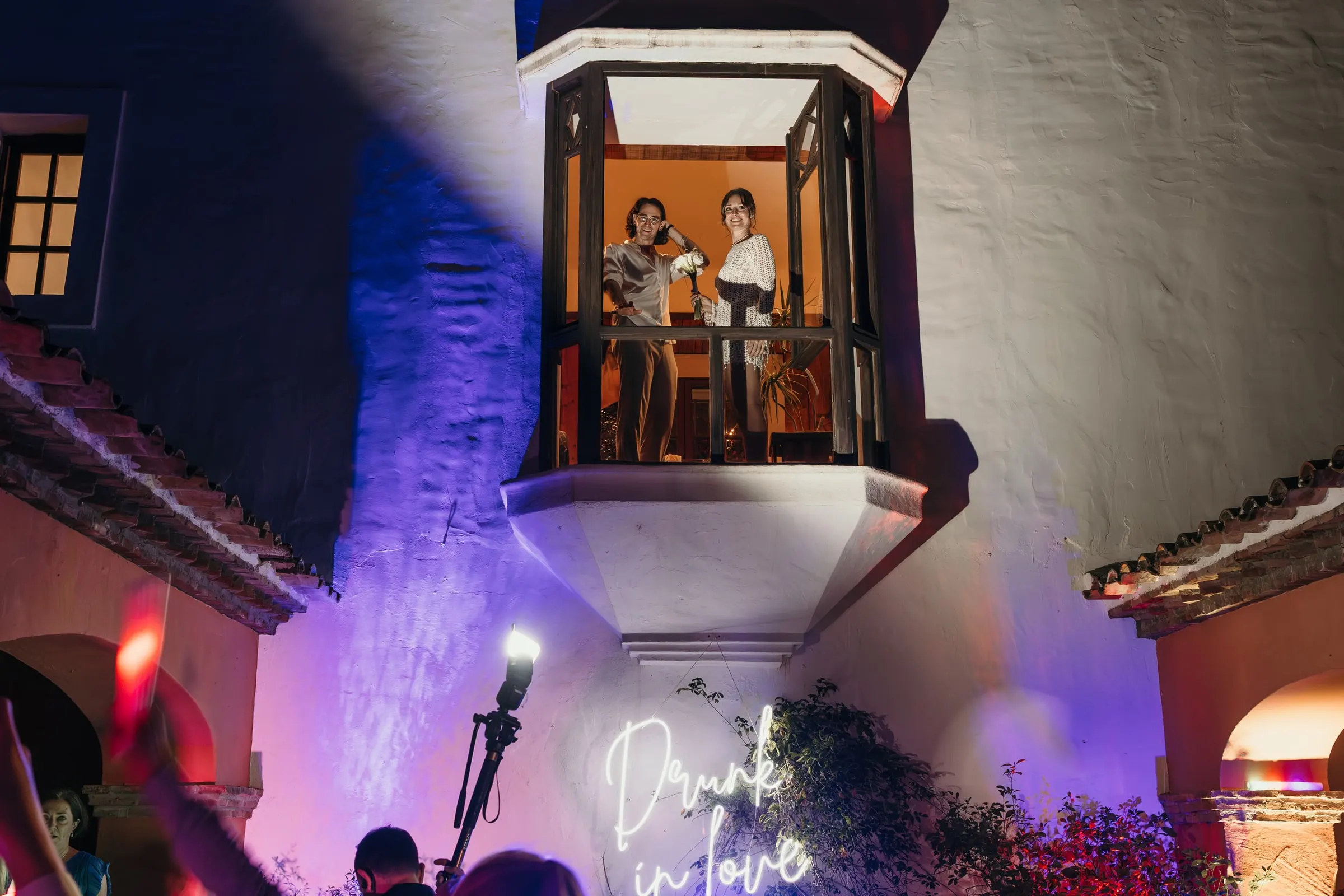 Bride and groom on a lit balcony above the party, with a neon sign and purple lights below, at Hacienda San José, Mijas