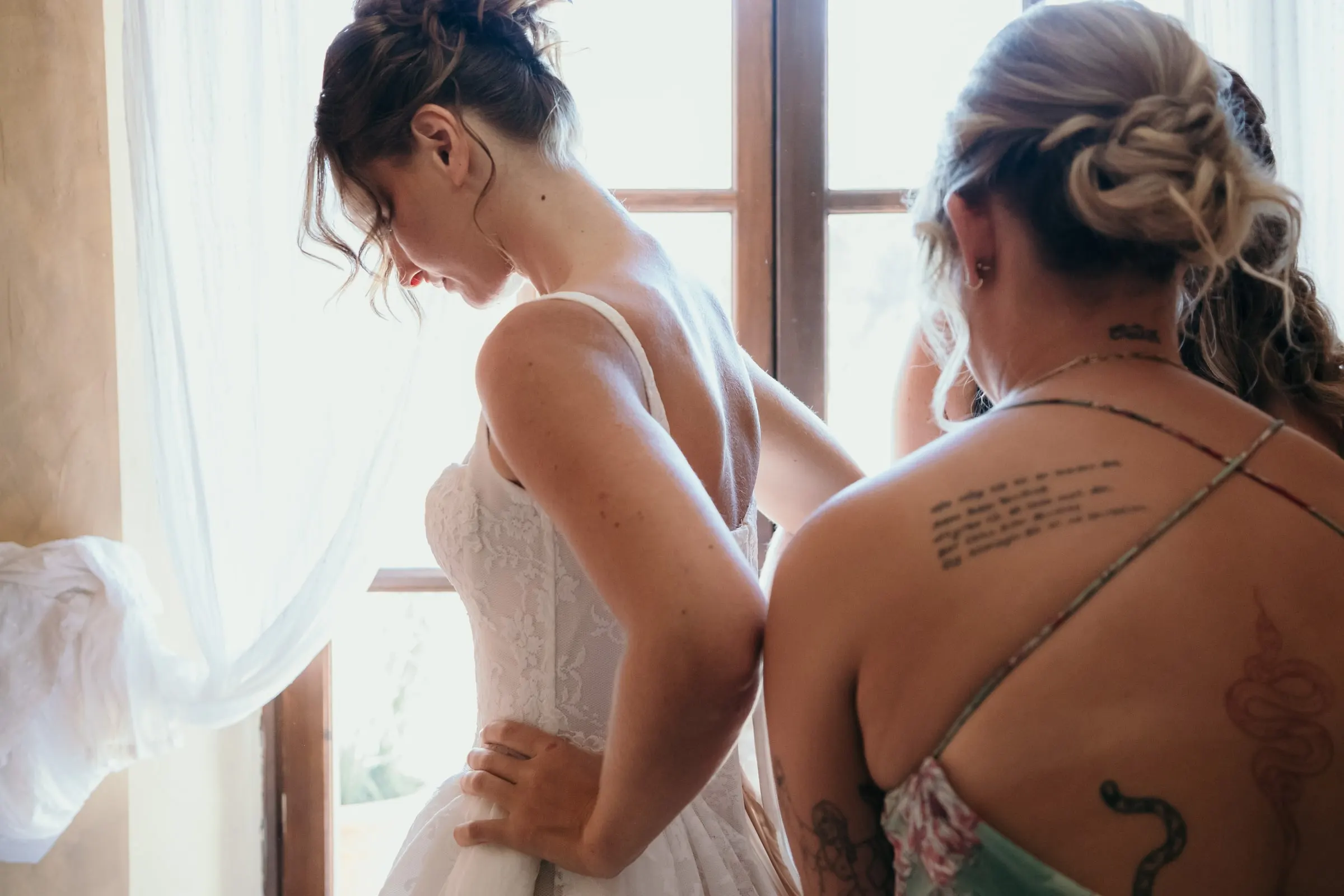 A tattooed friend helping the bride with her dress by the window, Hacienda San José, Mijas