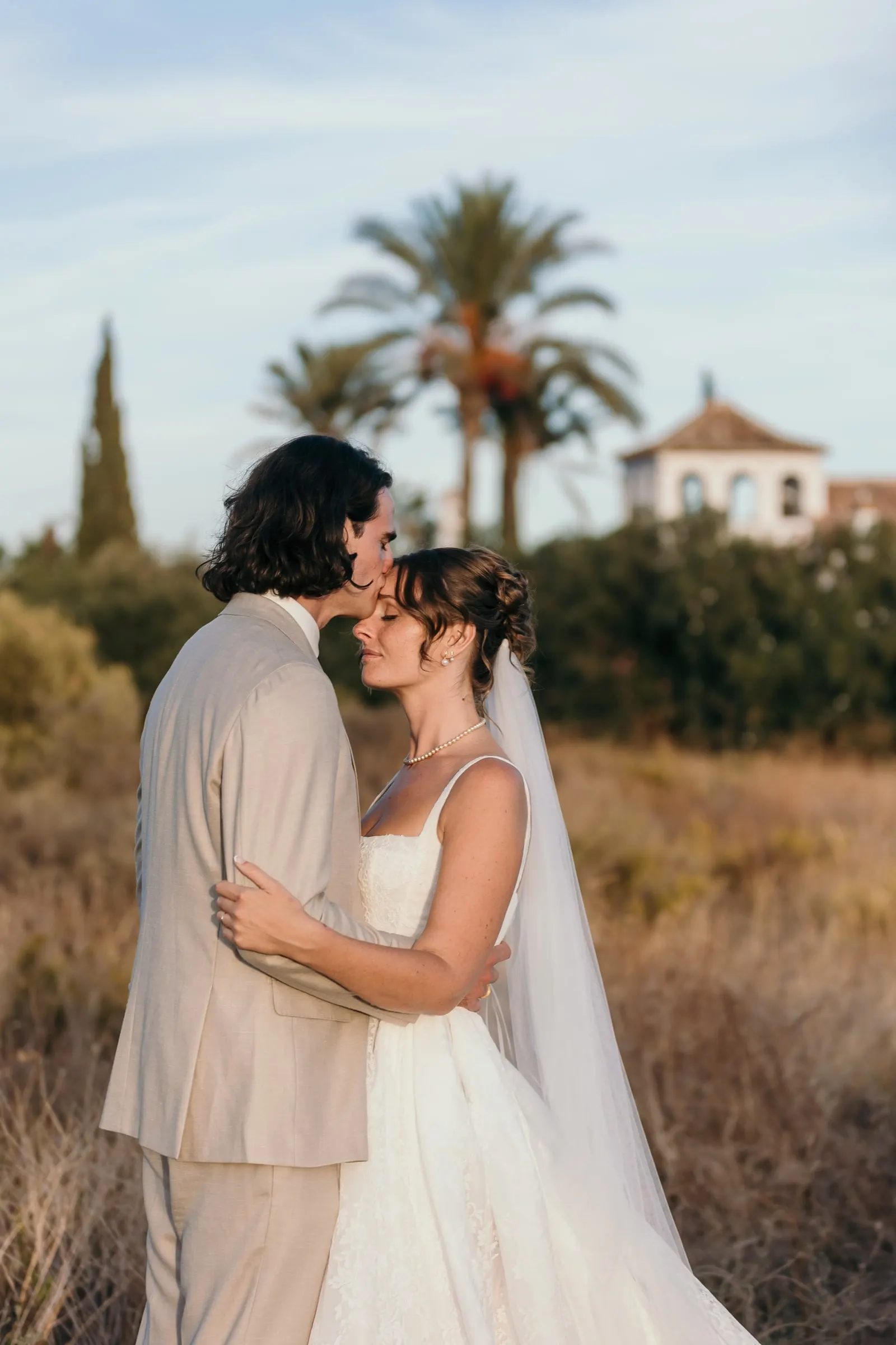 The groom kissing the bride's forehead in a sunlit field with palms behind, Hacienda San José, Mijas