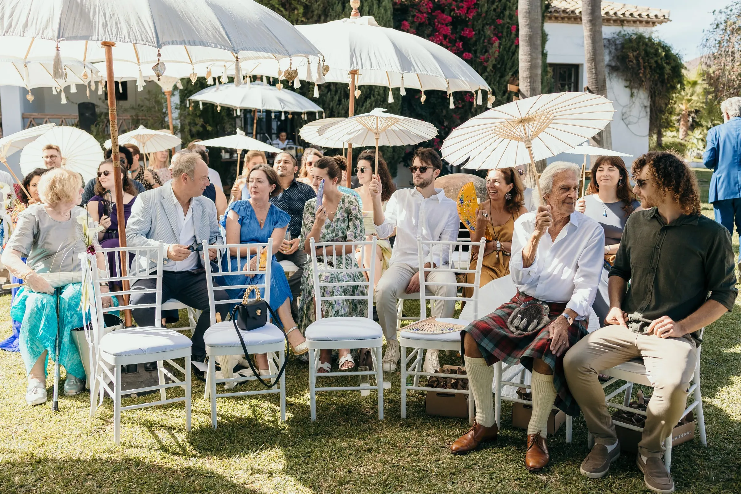 Guests seated under white parasols during the garden ceremony, one in a kilt, Hacienda San José, Mijas