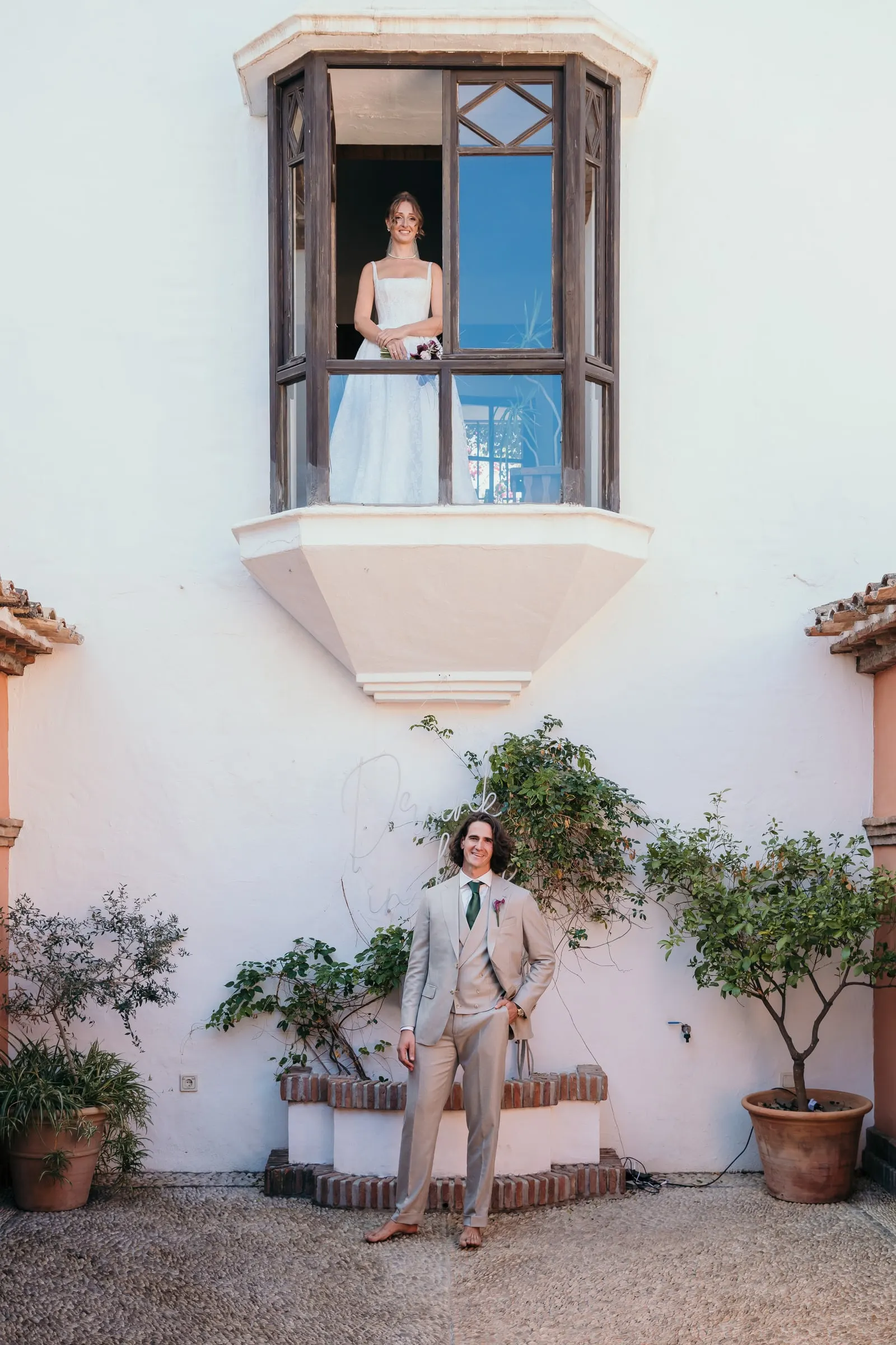 The bride at a balcony window above as the groom waits in the white courtyard, Hacienda San José, Mijas