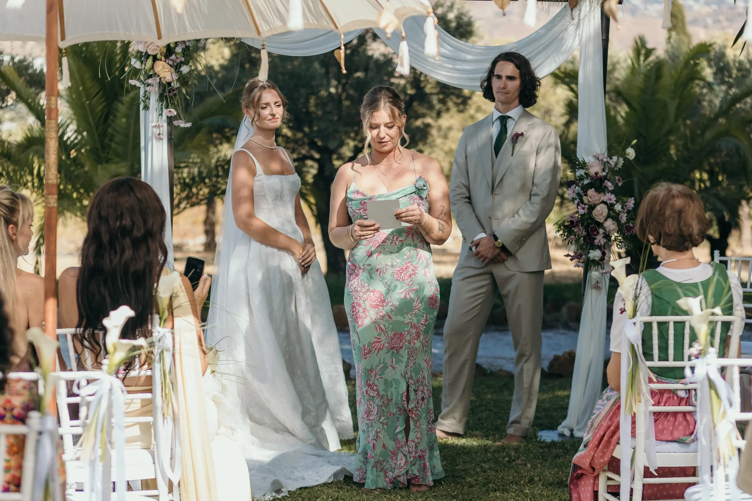 Bride and groom at the ceremony as a woman in a floral dress gives a reading, at Hacienda San José, Mijas