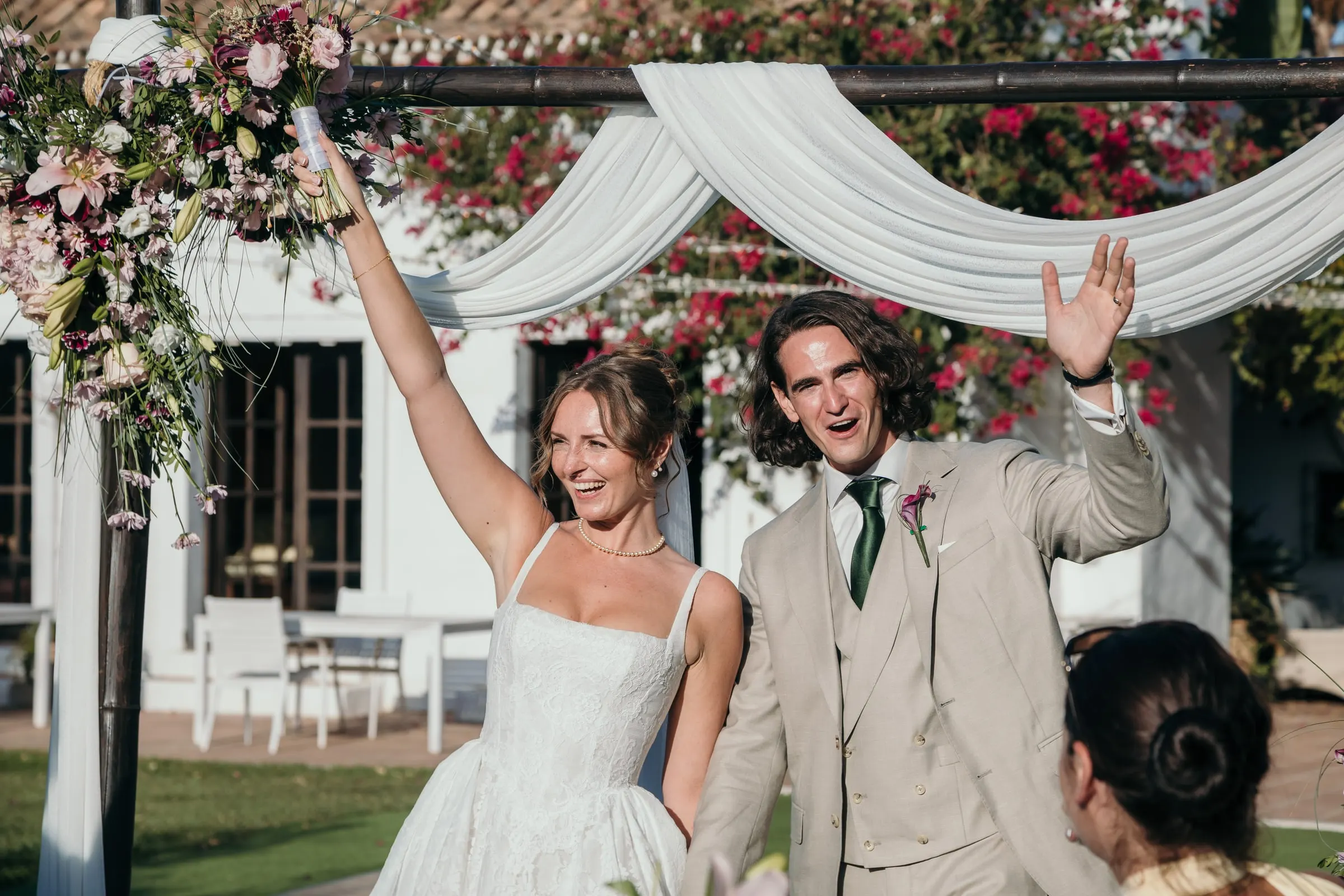 Bride and groom cheering with their arms raised under the floral arch after the ceremony at Hacienda San José, Mijas