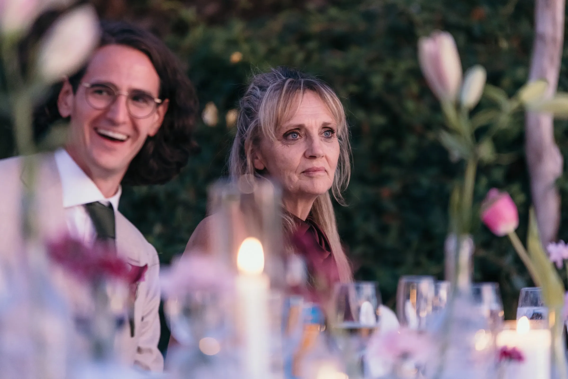 Groom laughing beside a guest at the dinner table, seen through pink flowers and candles, at Hacienda San José, Mijas