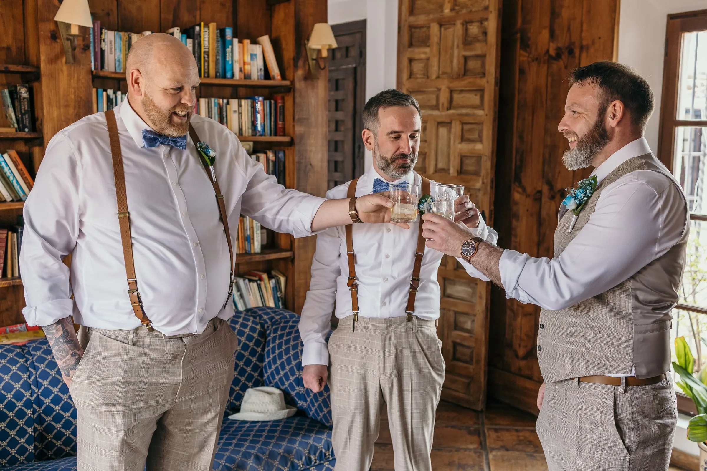 Groom and two friends in braces and blue bow ties raising a toast among the bookshelves in Mijas