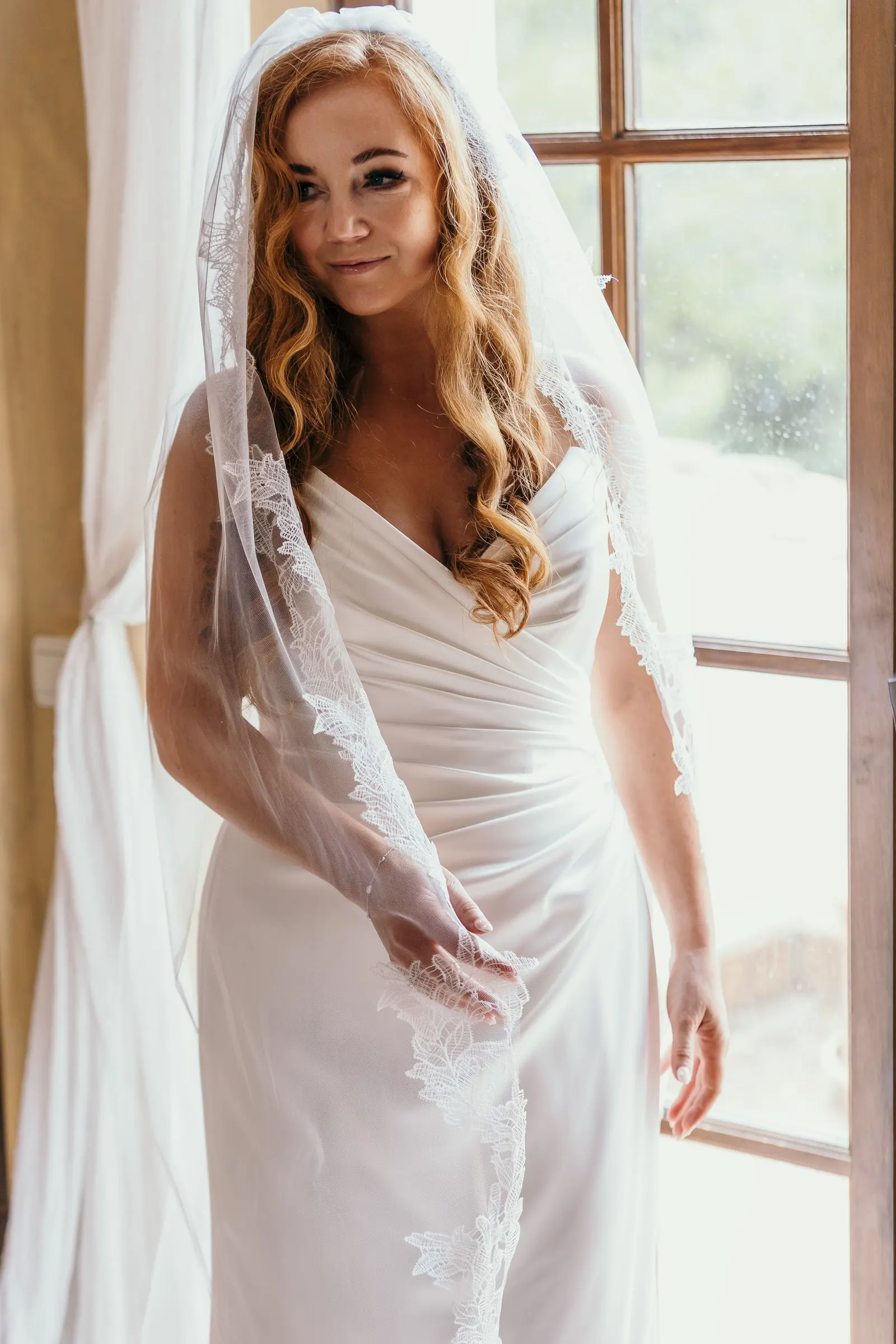 Bride in a lace-edged veil smiling by the tall window at Hacienda San José