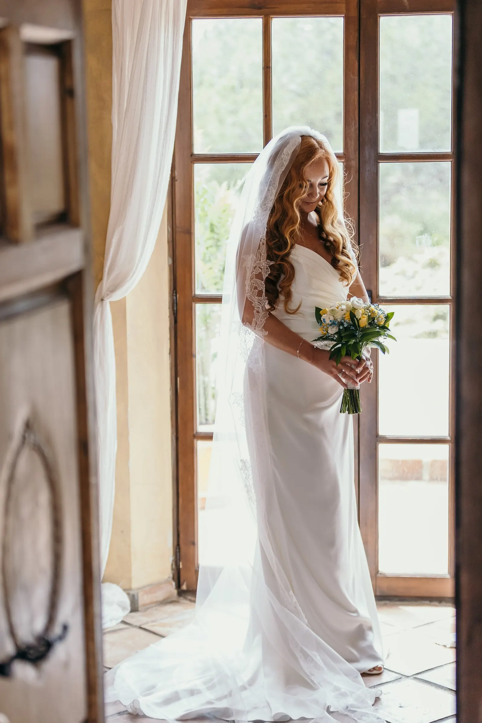Bride with her bouquet standing at the open French windows, long train on the tiles, Mijas