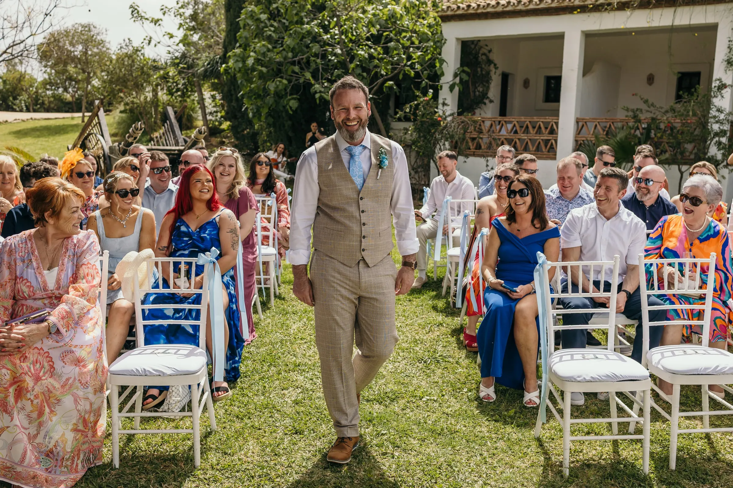Groom in a beige waistcoat walking down the garden aisle between the guests at Hacienda San José