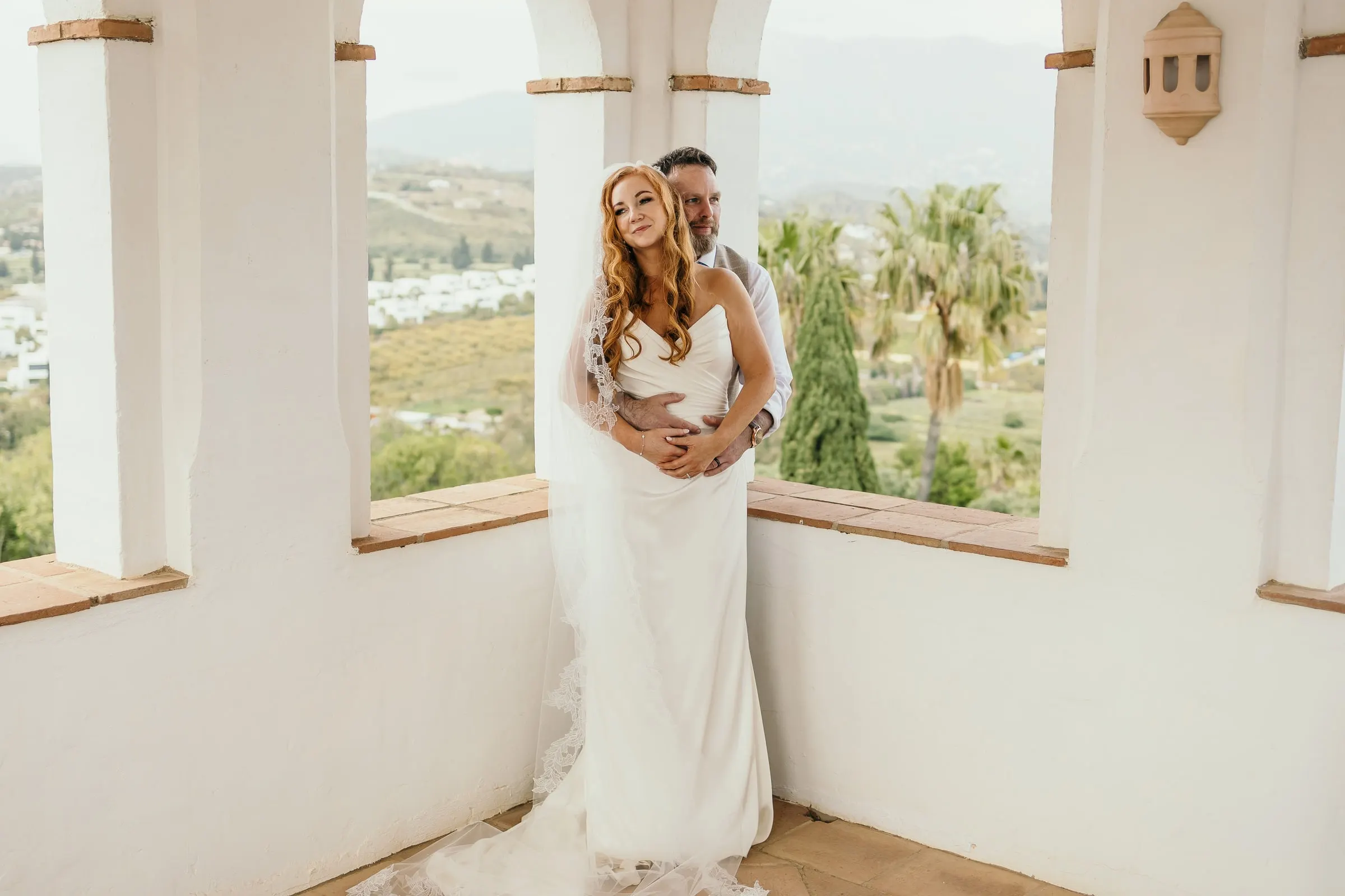 Groom embracing the bride from behind in the tower loggia, palm trees and hills beyond, Mijas