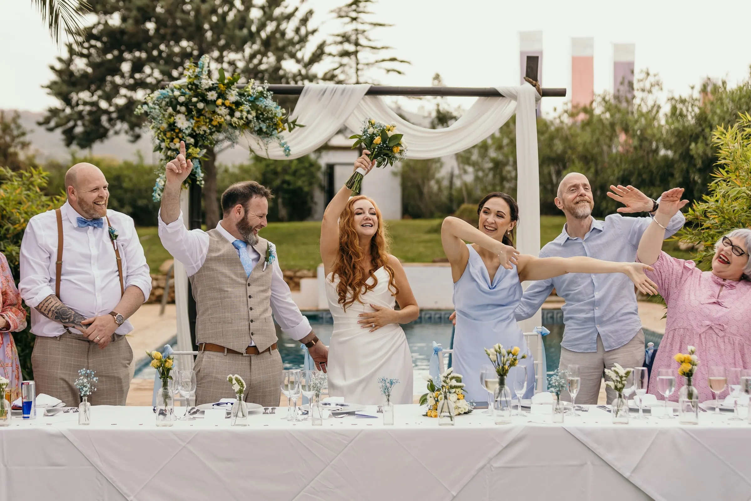 Wedding party cheering and raising their arms at the top table by the pool, Hacienda San José