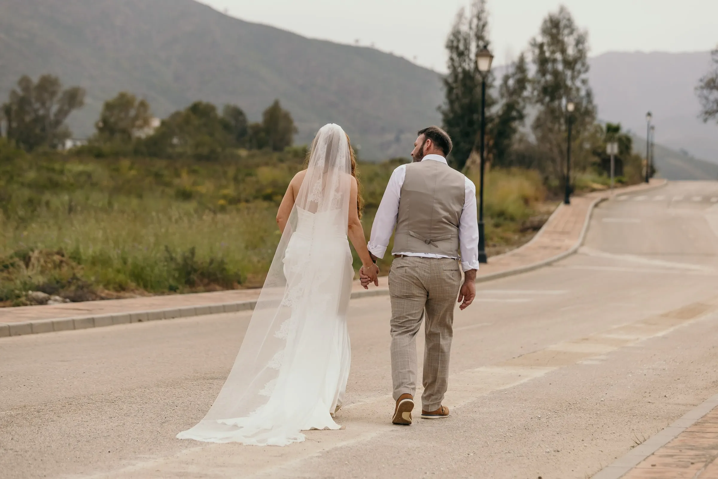 Bride and groom walking hand in hand down a quiet road into the hills at sunset, Mijas