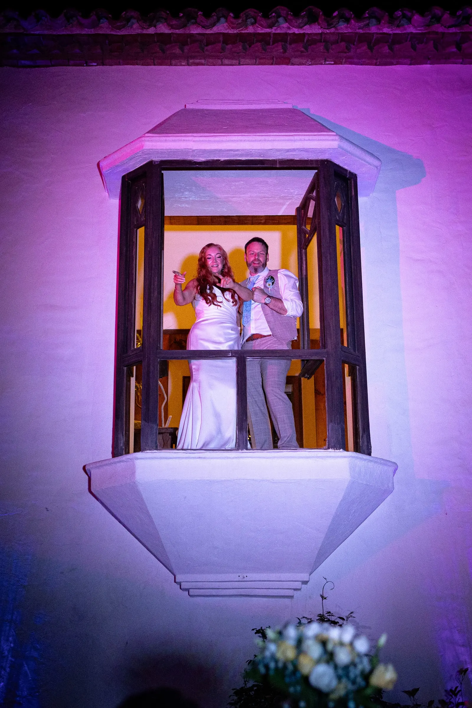 Couple waving from the window bay above the guests at night, Hacienda San José