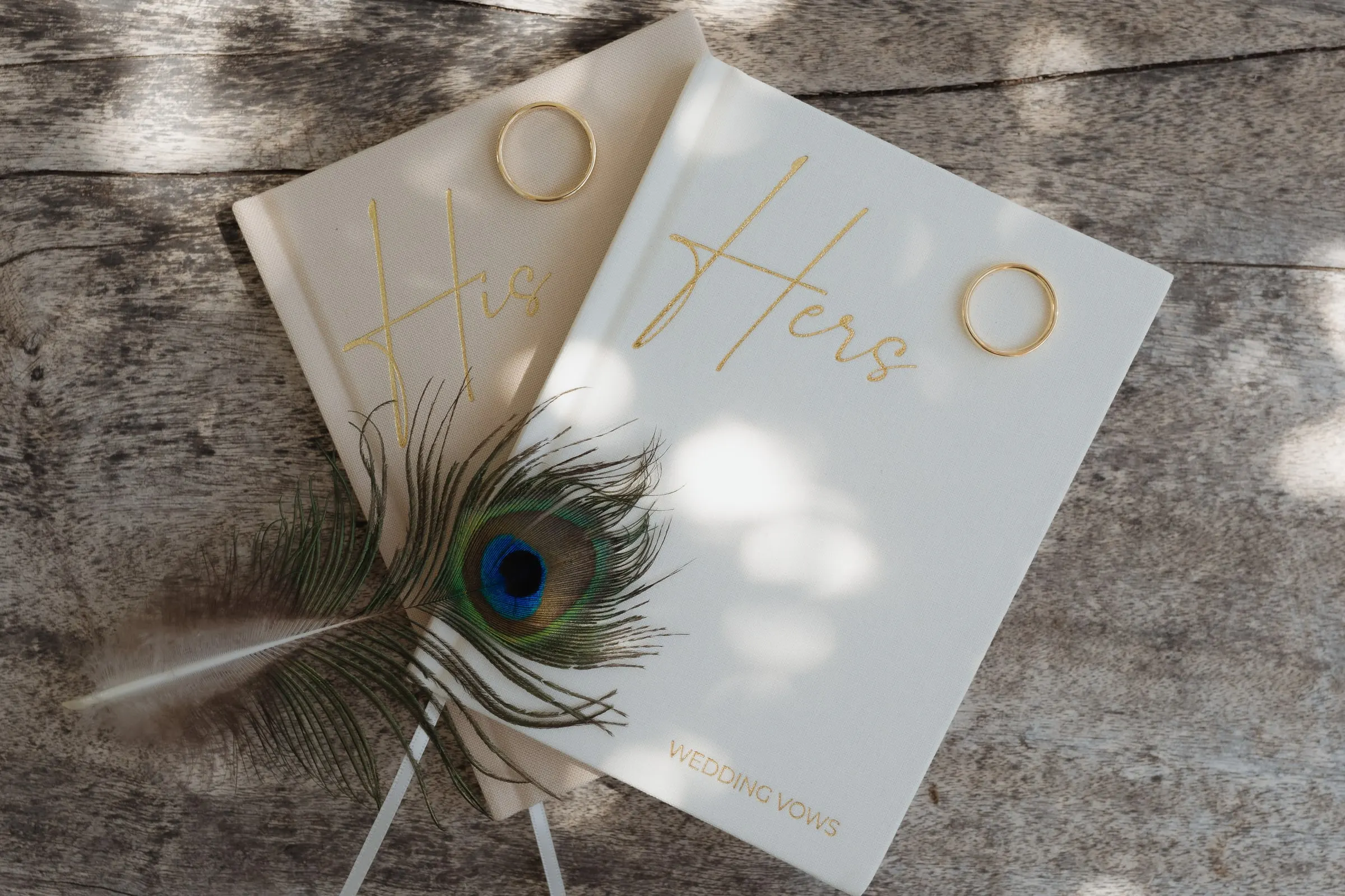 Libros de votos «His» y «Hers» con una pluma de pavo real sobre madera envejecida