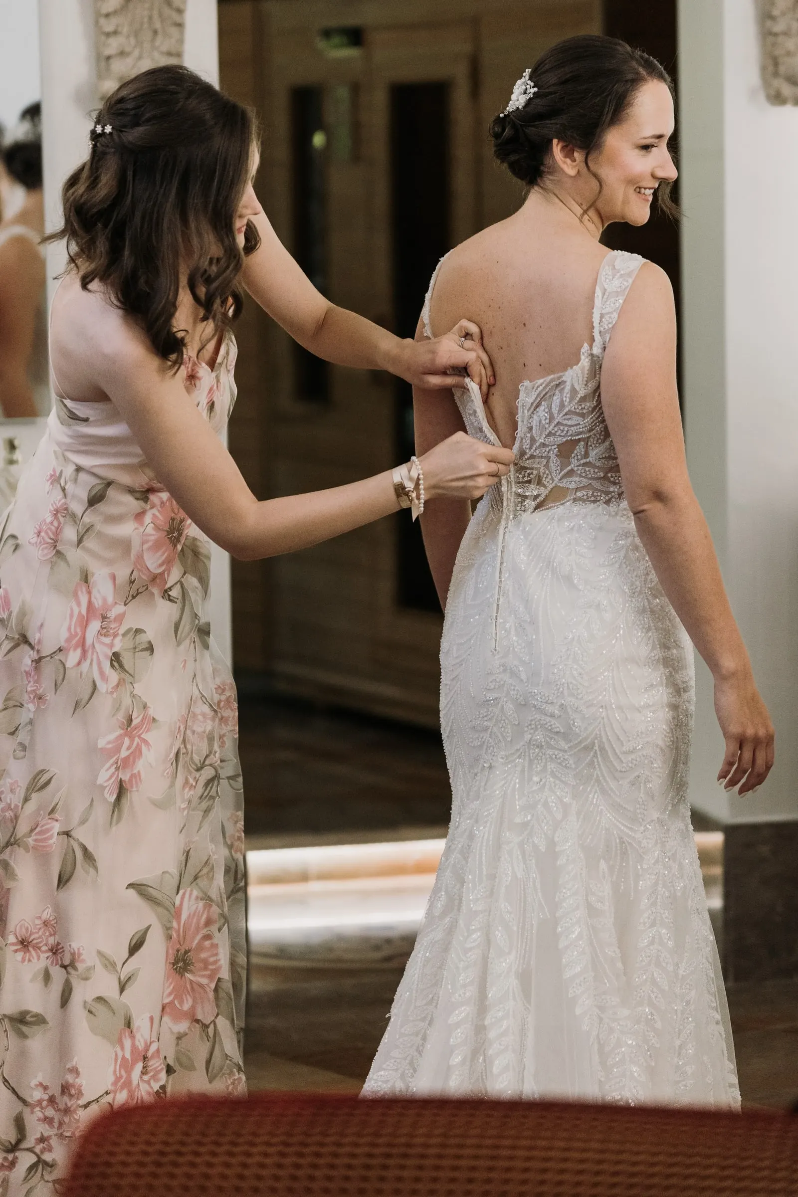 Una amiga con vestido de flores abrocha la espalda del vestido de encaje de la novia