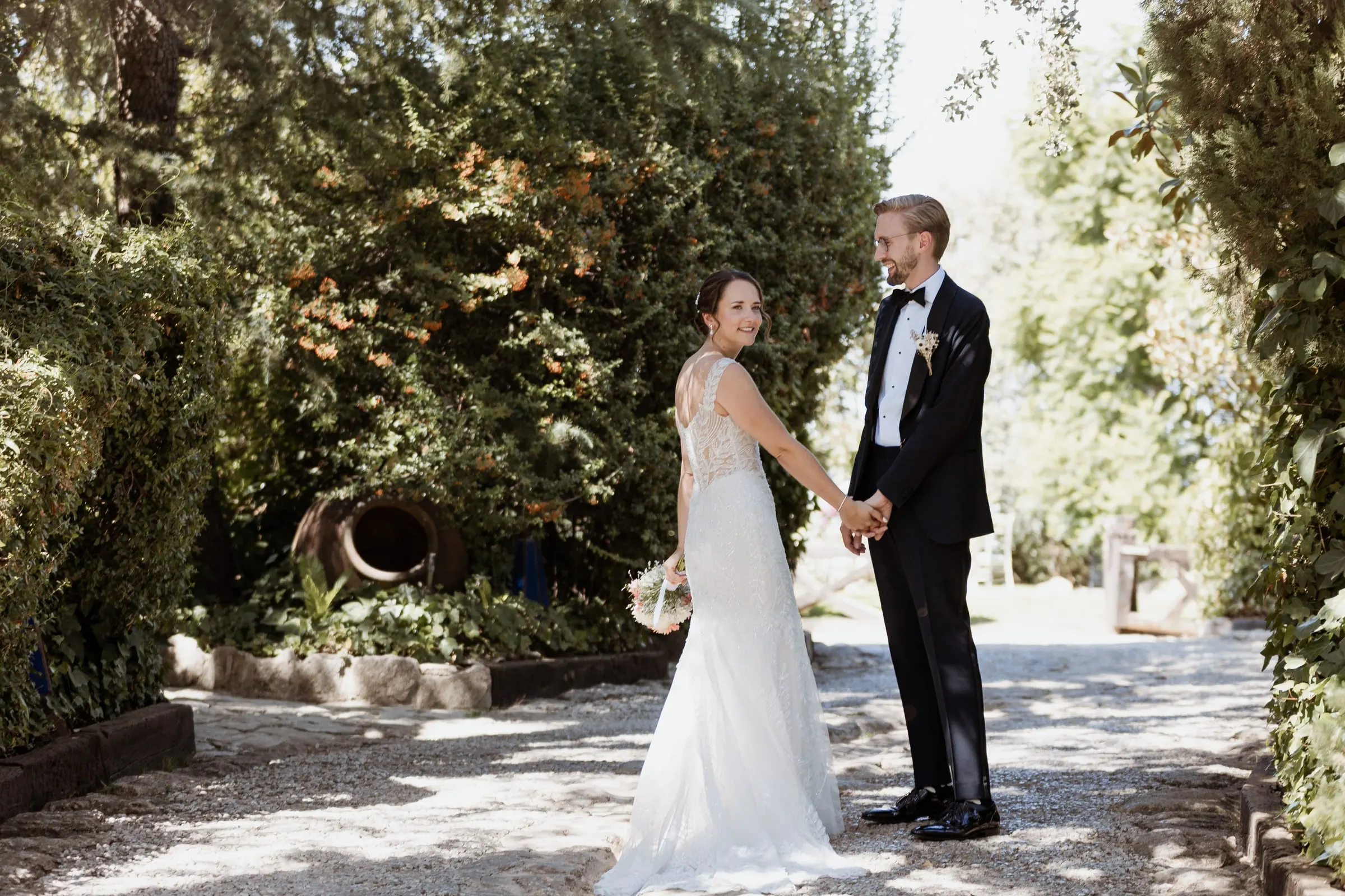 Los novios de la mano en un camino de grava bajo un túnel de árboles