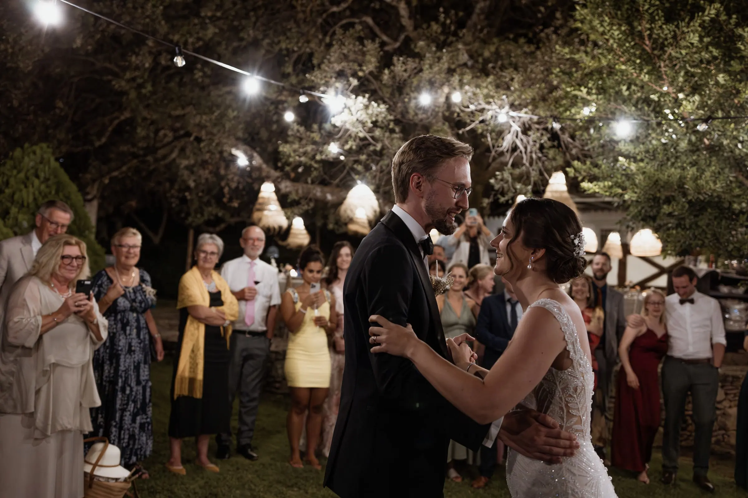 La pareja baila rodeada de invitados bajo los árboles de noche