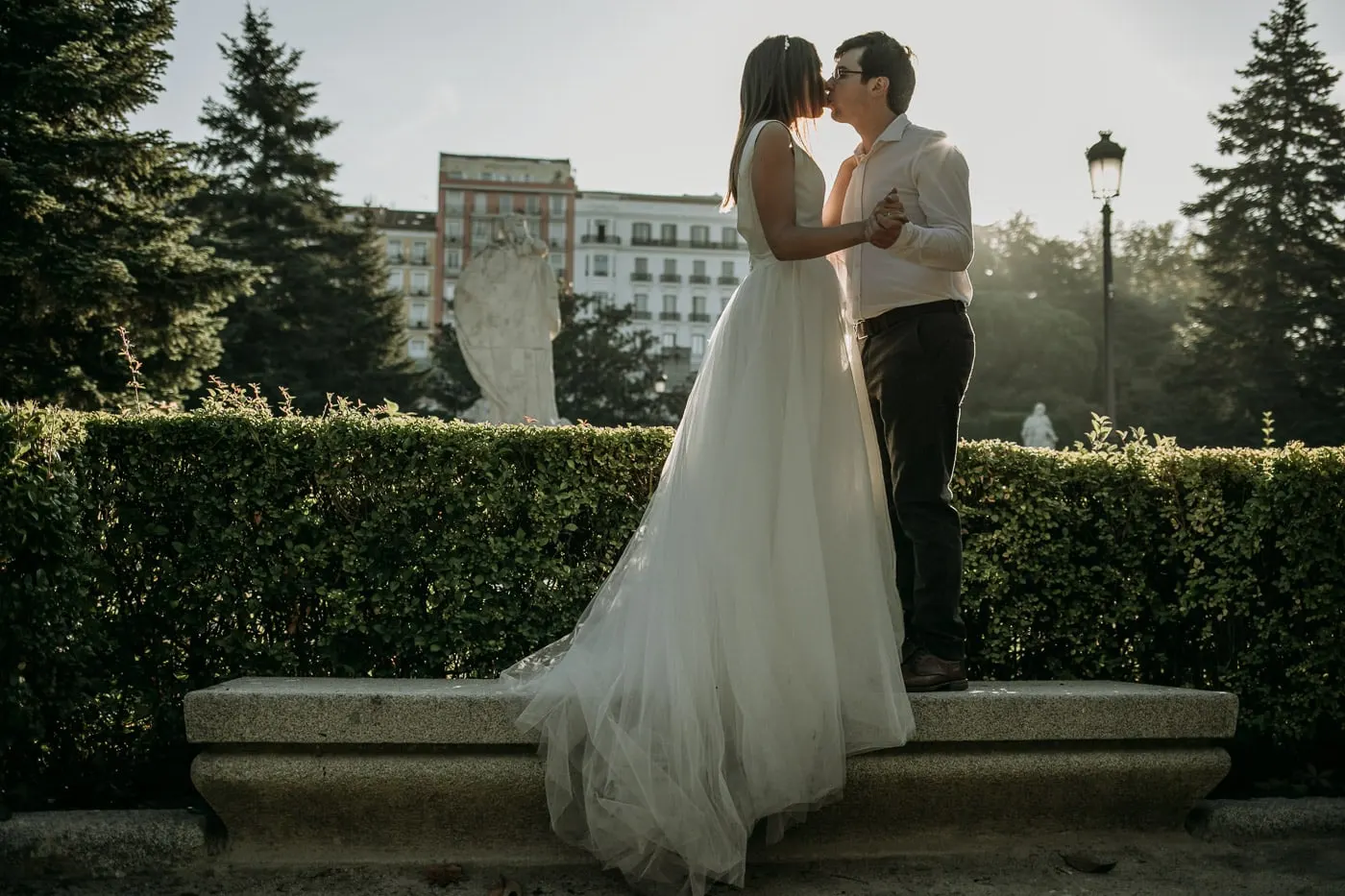 La novia, subida a un banco de piedra, besa al novio en los jardines del palacio