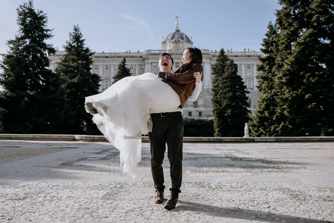 El novio lleva en brazos a la novia y se ríe frente al Palacio Real, Madrid