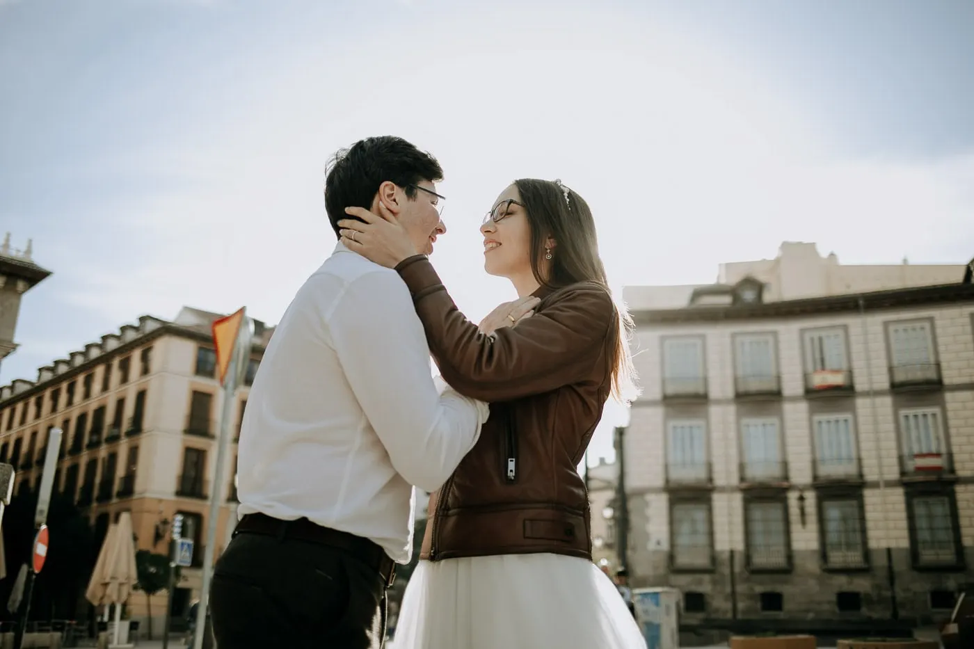 El novio sostiene la cara de la novia contra el cielo claro, con edificios antiguos de Madrid detrás
