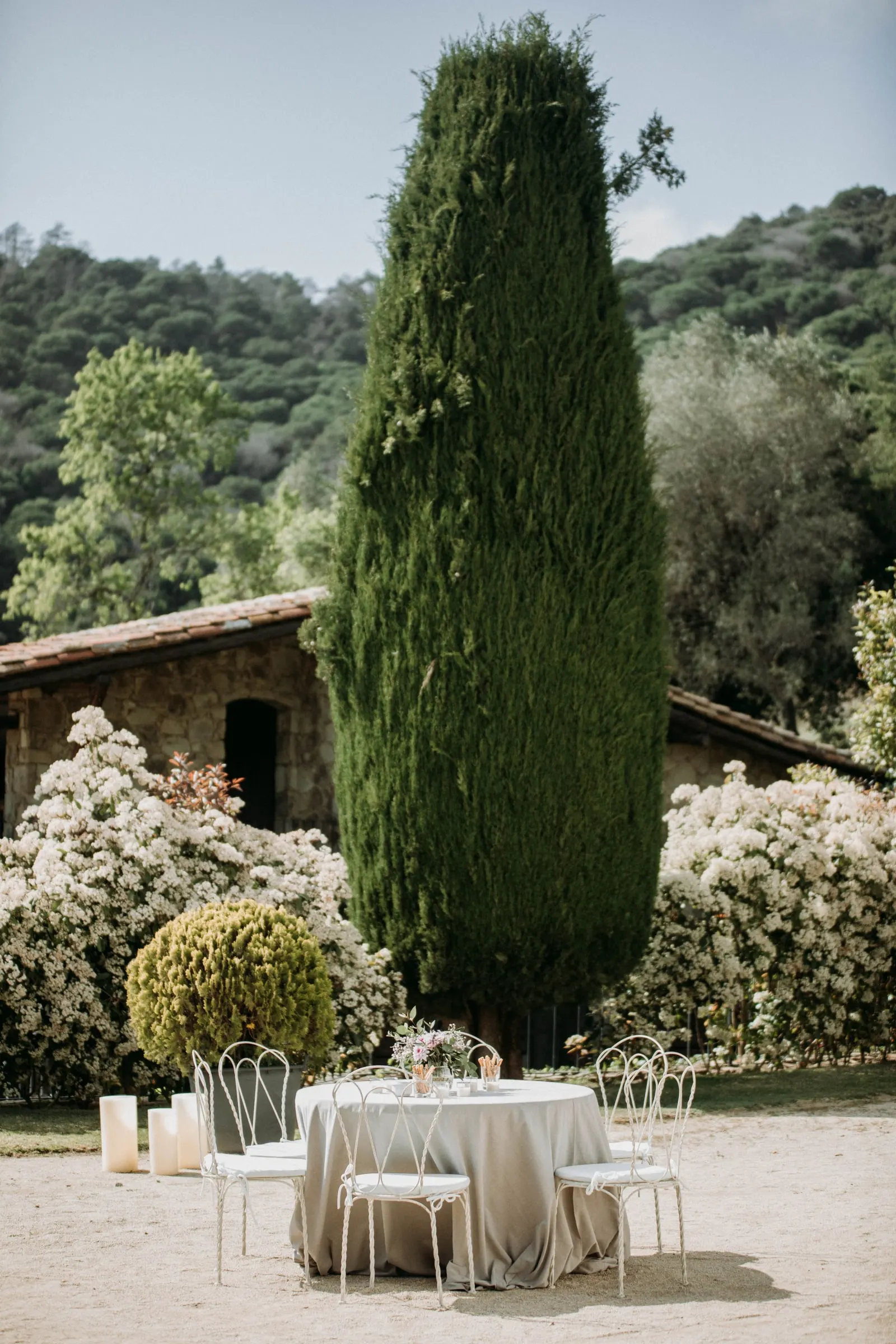 Una mesa para dos junto a un ciprés y arbustos en flor en el jardín de una masía de Girona