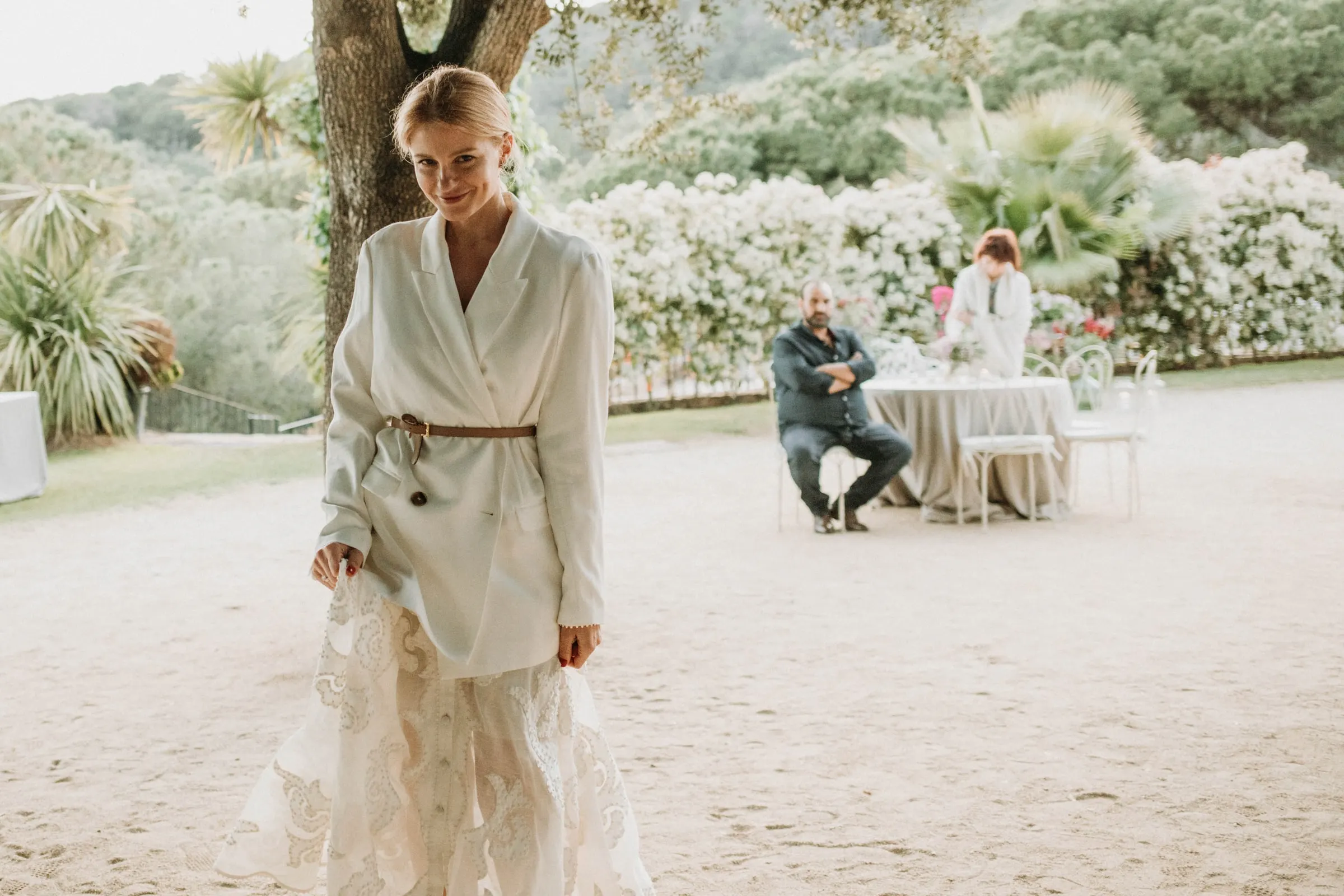 La novia, con americana blanca y falda bordada, sonríe en el jardín de grava de una masía de Girona