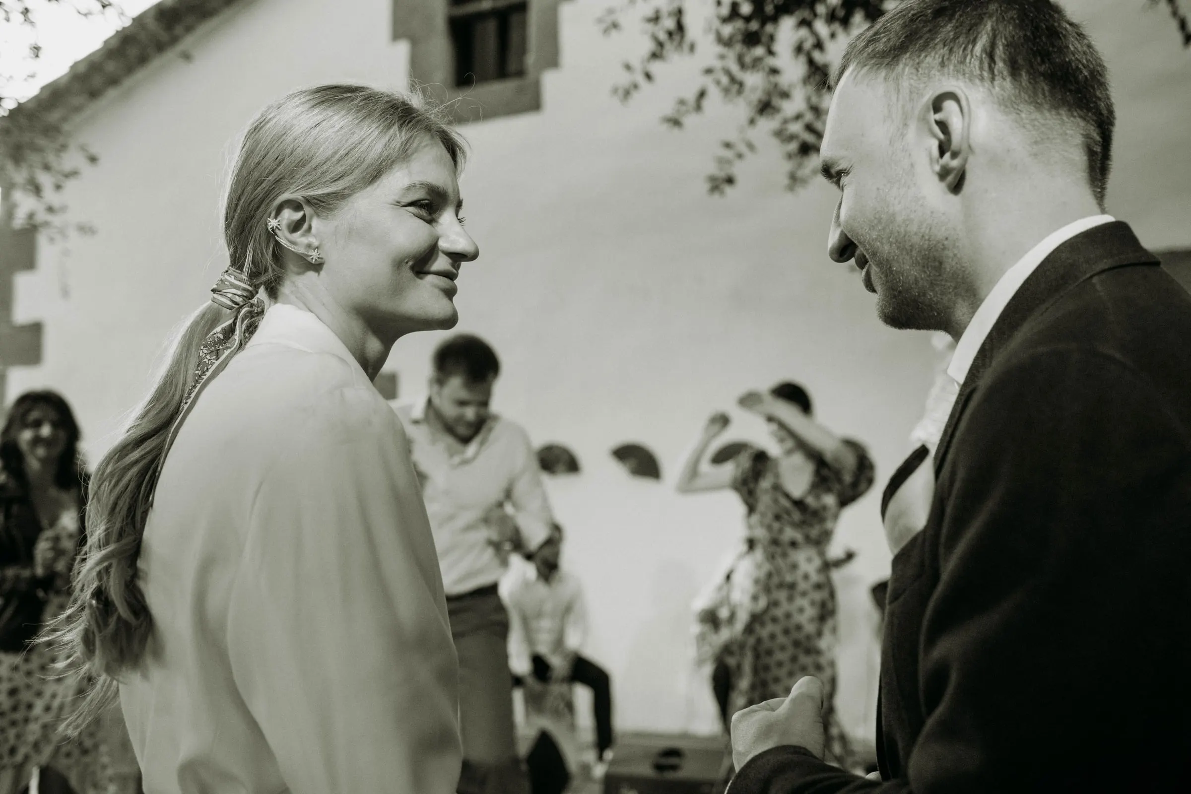 Los novios se sonríen en blanco y negro mientras los invitados bailan detrás, en una masía de Girona