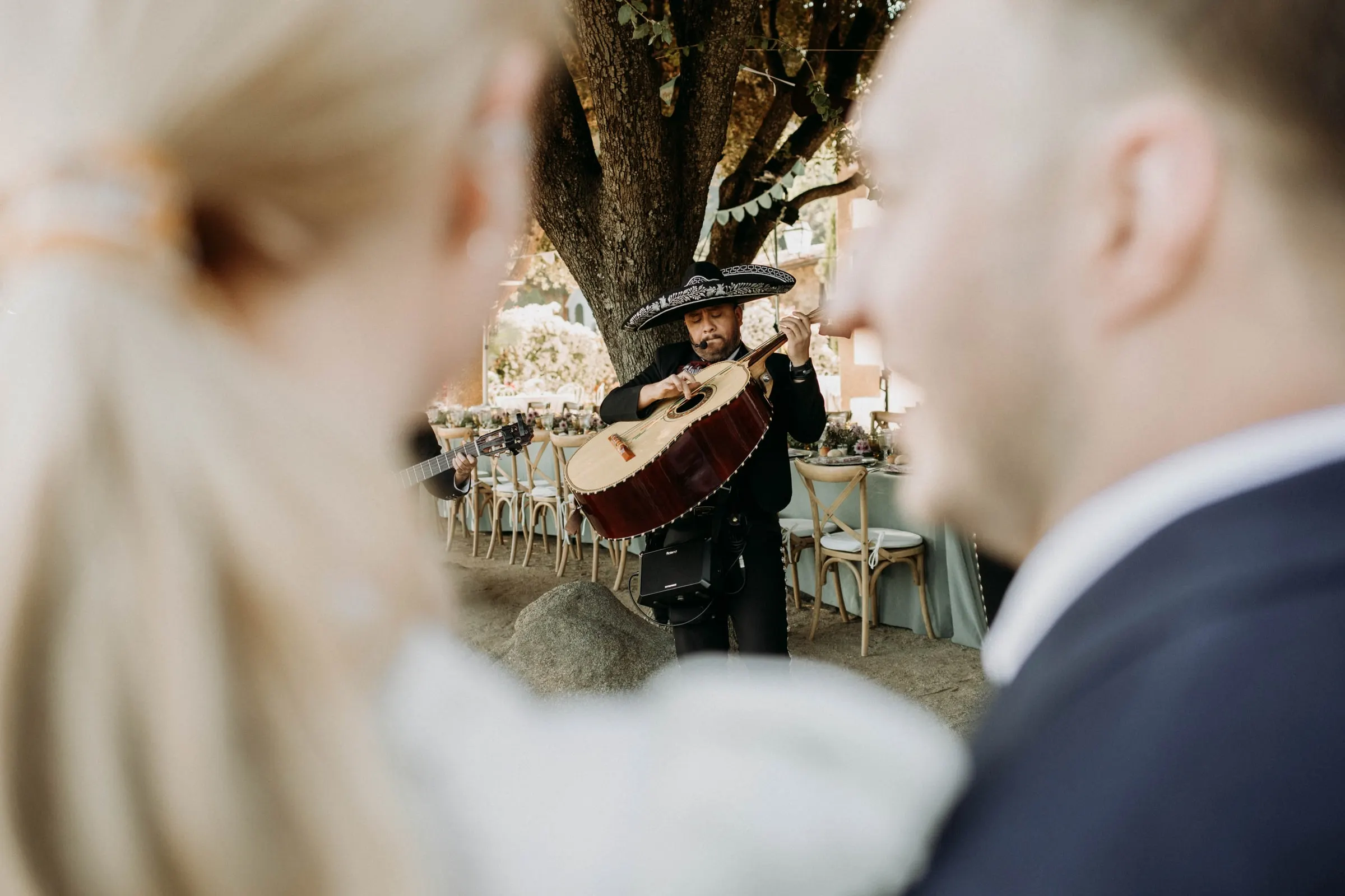 Un mariachi toca el guitarrón bajo un árbol, visto entre los novios, en una masía de Girona