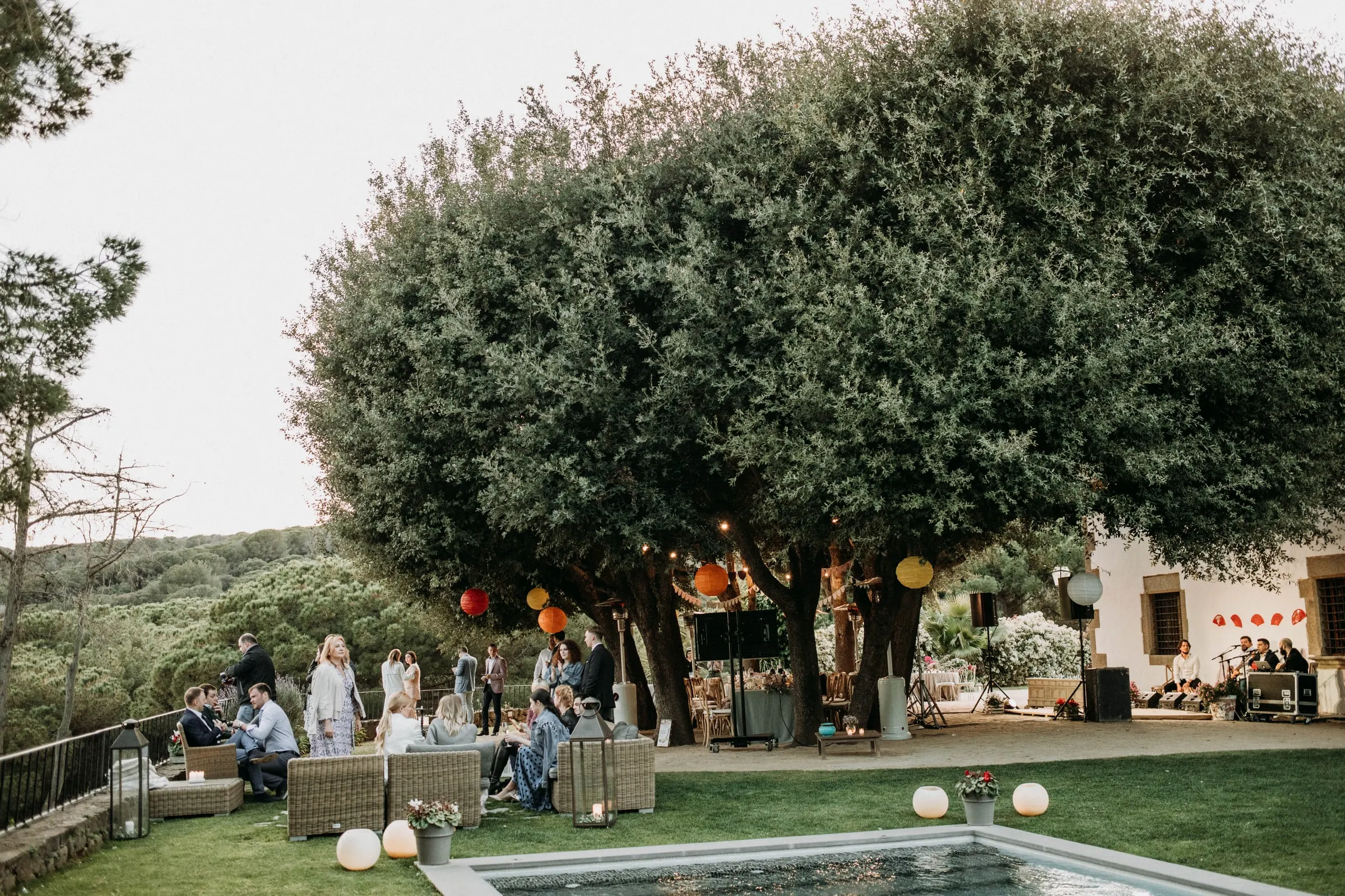 Invitados en los sofás del jardín junto a la piscina, bajo un gran árbol con farolillos, en una masía de Girona