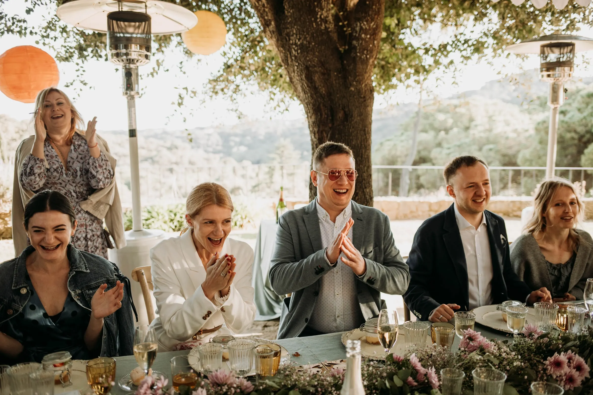 La novia y los invitados aplauden y ríen en la mesa principal bajo el árbol, en una masía de Girona