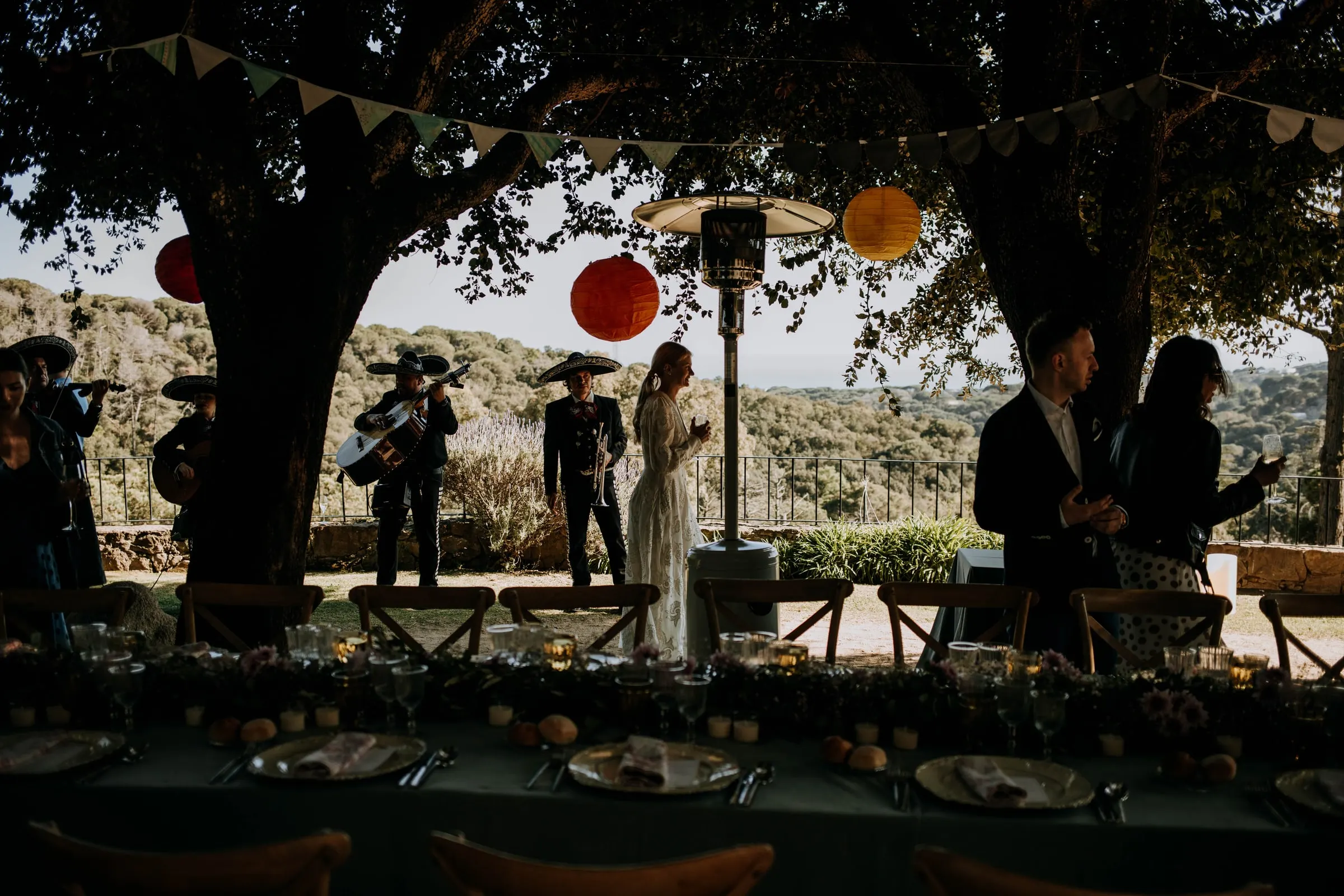 Los mariachis dan una serenata a los novios al fondo de la mesa larga bajo los árboles, en una masía de Girona